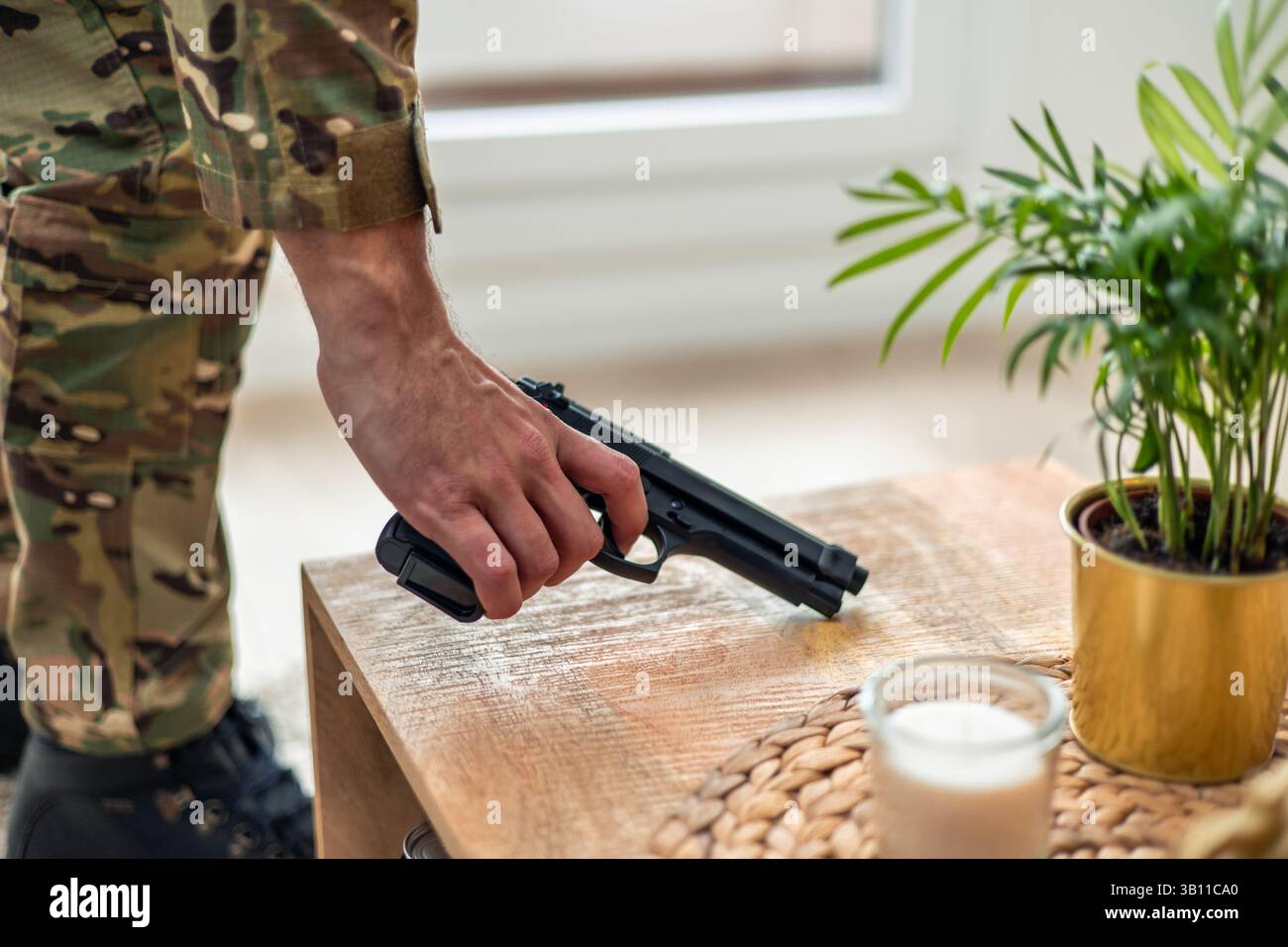 Soldato in uniforme mimetica che prende una pistola da un tavolo di legno a casa, lottando contro il disturbo post-traumatico dello stress e il suo impatto sulla salute mentale Foto Stock