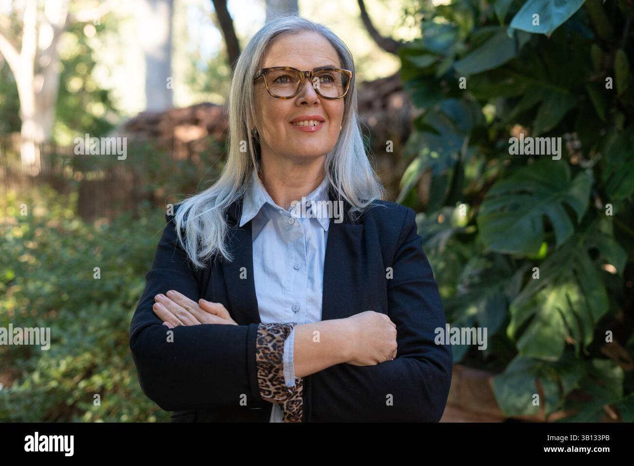 Donna d'affari con le braccia incrociate, sorridente mentre guarda lontano, si posa in un tranquillo parco circondato da lussureggianti piante verdi, incarnando leader Foto Stock