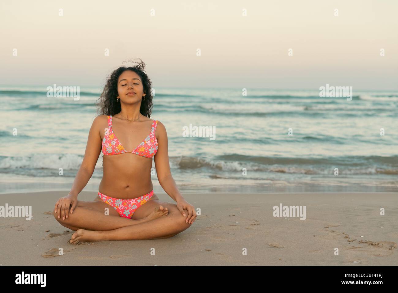 Donna in un bikini floreale che medita su una spiaggia tranquilla al tramonto, abbracciando il relax e la consapevolezza accanto alle onde dell'oceano. Atmosfera tranquilla e serena Foto Stock