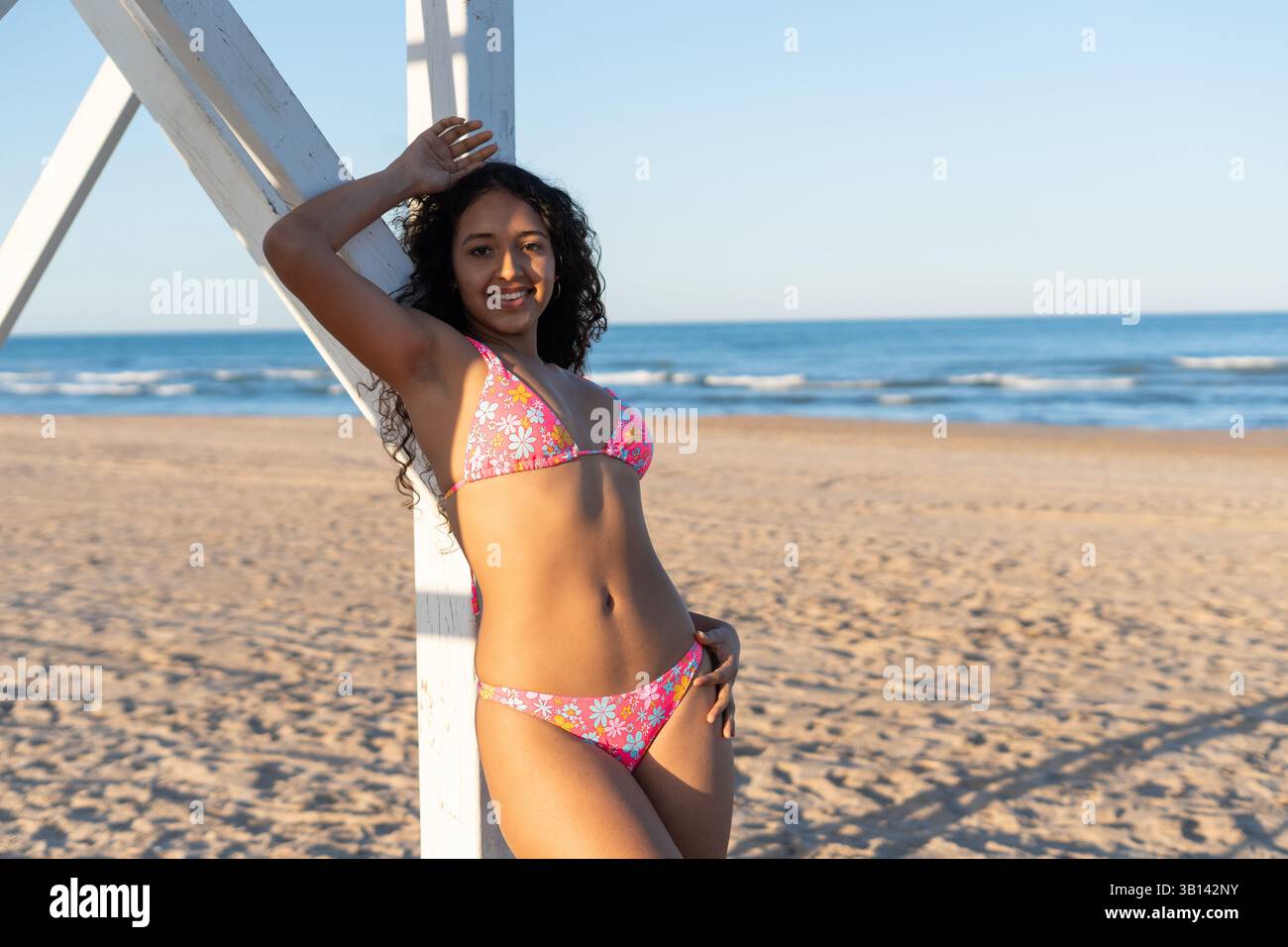 Donna sorridente in un bikini colorato che si gode una giornata di sole sulla spiaggia, appoggiata a un bagnino con l'oceano sullo sfondo. Scen perfetto Foto Stock