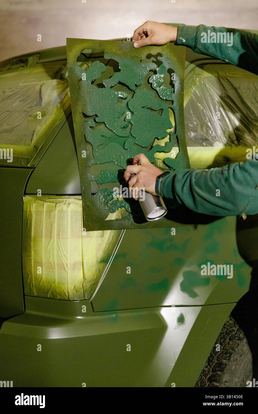 Una persona applica un design mimetico verde su un veicolo utilizzando una bomboletta spray e uno stencil. L'allestimento dell'officina mostra i preparativi per la verniciatura, Highlig Foto Stock
