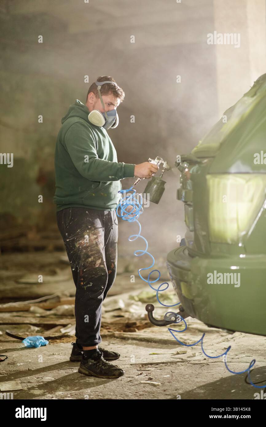 Un uomo che indossa una maschera e un equipaggiamento protettivo si concentra sulla verniciatura di un'auto all'interno di un'officina disordinata. Polvere e detriti lo circondano mentre lavora meticoloso Foto Stock
