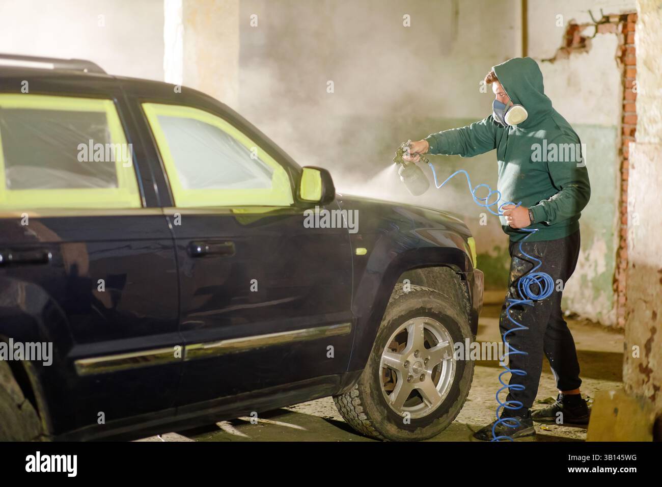Una persona che indossa una giacca con cappuccio e una maschera sta dipingendo un'auto di colore scuro in un garage. La luce soffusa e la nebbia creano un'atmosfera mirata per il Foto Stock