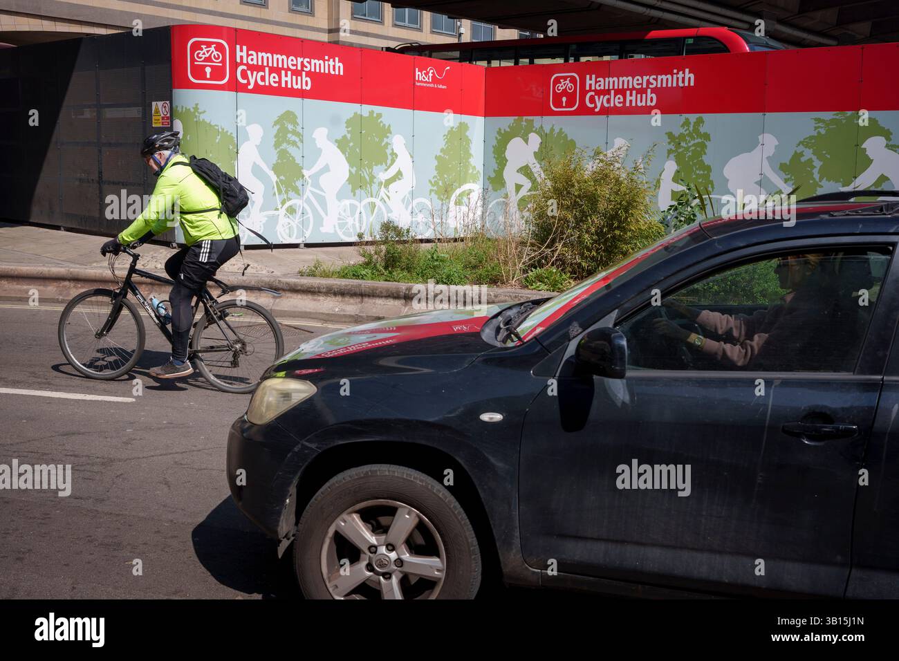 Un ciclista passa un'auto e un cartellone per l'Hammersmith Cycle Hub, il 24 aprile 2025, a Londra, Inghilterra. I residenti del quartiere londinese di Hammersmith & Fulham e i pendolari possono parcheggiare le loro biciclette in modo sicuro presso l'Hammersmith Cycle Hub, situato sotto l'Hammersmith Flyover in cima a Fulham Palace Road, fornendo una soluzione per i viaggiatori che desiderano andare in bicicletta alla stazione di Hammersmith, lasciare la bicicletta e continuare il viaggio in metropolitana o autobus. Foto Stock