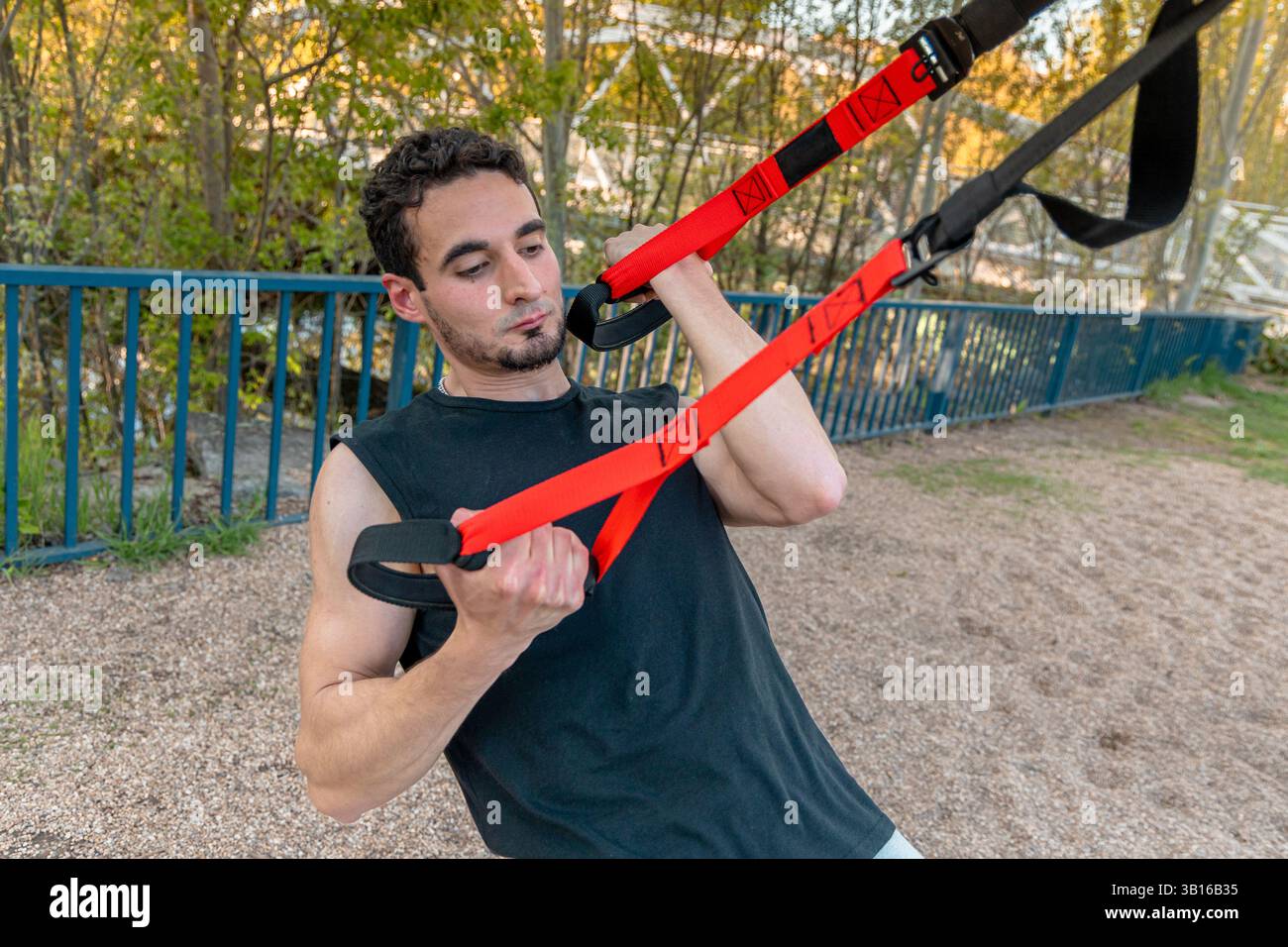 Giovane uomo che esegue sospensioni TRX all'aperto utilizzando cinghie rosse, concentrandosi su forza, muscoli della schiena e controllo durante l'allenamento con il peso corporeo Foto Stock