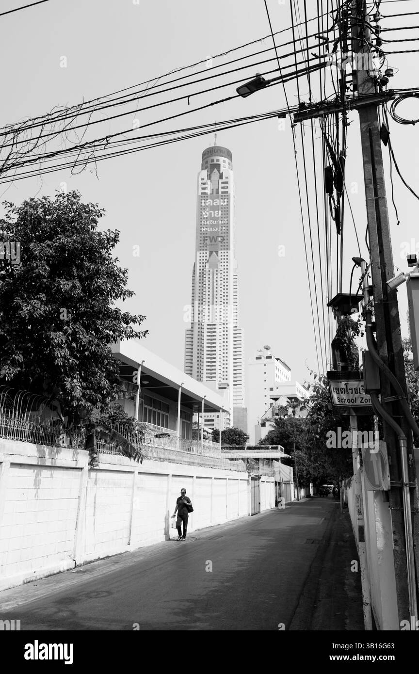 Un solo pedonale cammina lungo un vicolo ombroso sotto la torreggiante Baiyoke Tower II Foto Stock