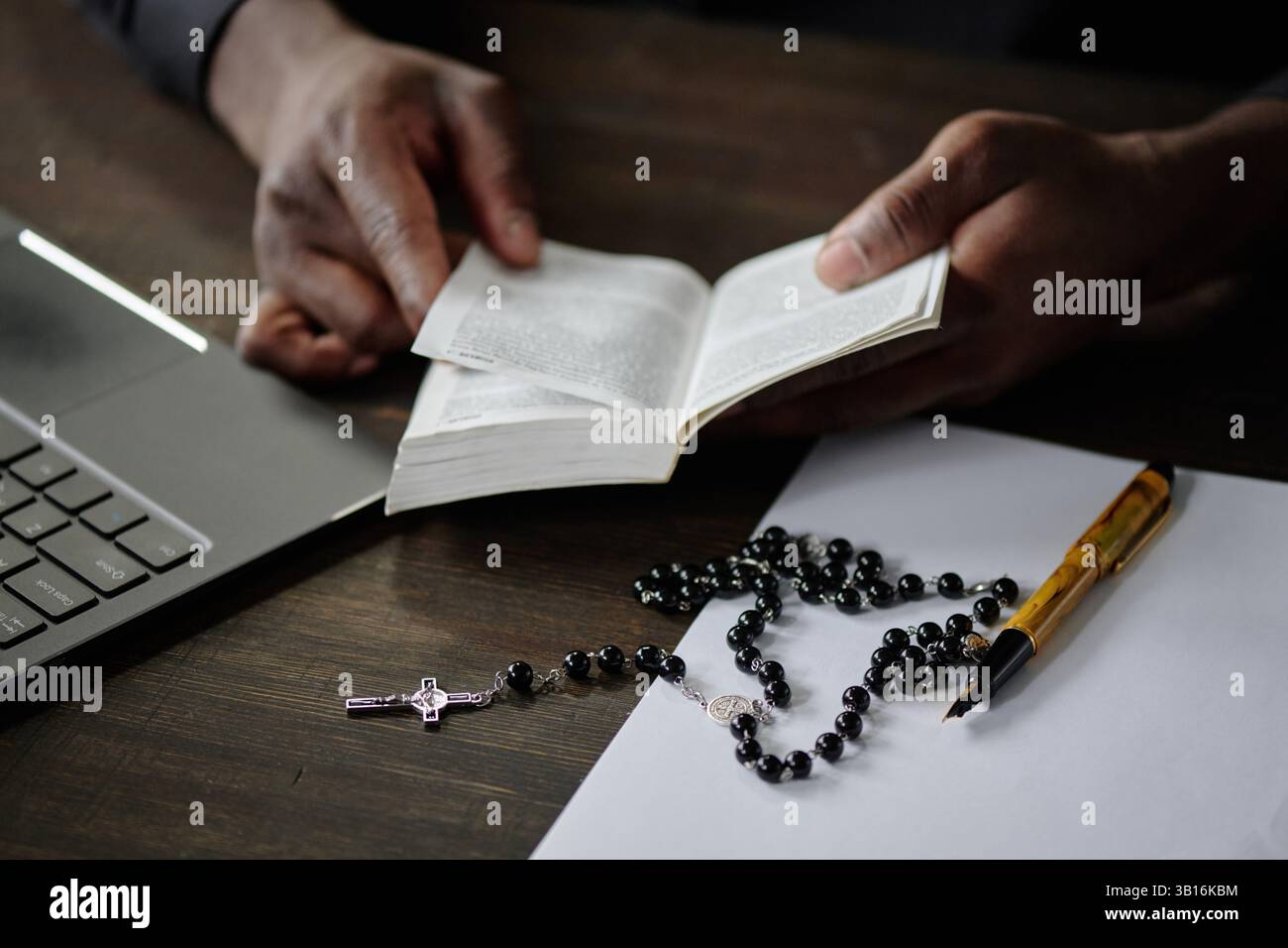 Medio primo piano di pastori irriconoscibili mani che tengono aperta la bibbia. Sedeva alla scrivania di legno con computer portatili, penna vintage e rosario con perle nere e croce Foto Stock