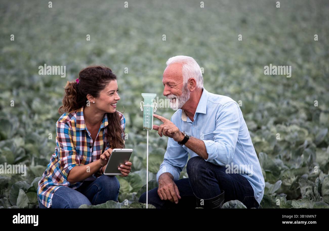 Due agricoltori stanno esaminando l'umidità del suolo nel pluviometro mentre sono circondati da file di colture in un campo soleggiato. Sono concentrati sul loro compito insieme Foto Stock