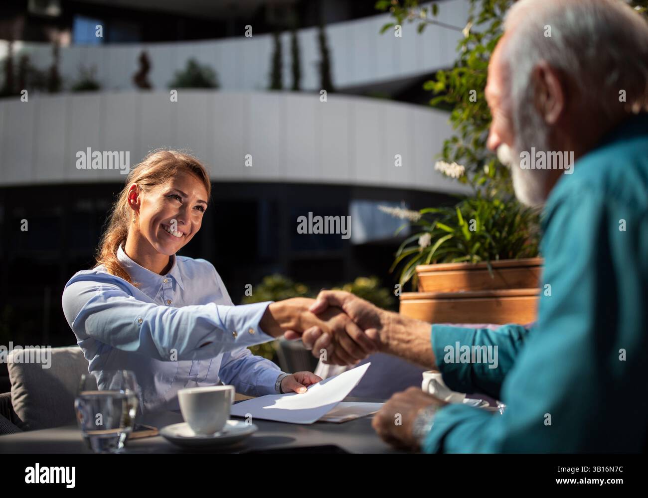 Giovane donna che stringe la mano a un uomo anziano in un bar all'aperto, entrambi vestiti professionalmente. Sembrano impegnati in una discussione d'affari sul caffè. Foto Stock