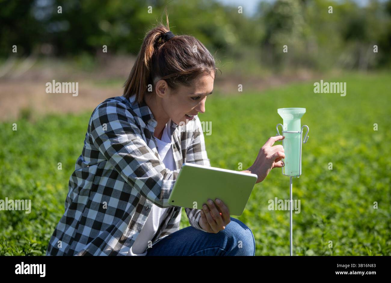Donna che controlla i dati su un tablet mentre osserva il pluviometro e le piante in un lussureggiante campo verde. L'attività si svolge sotto i cieli limpidi, mostrando rura Foto Stock