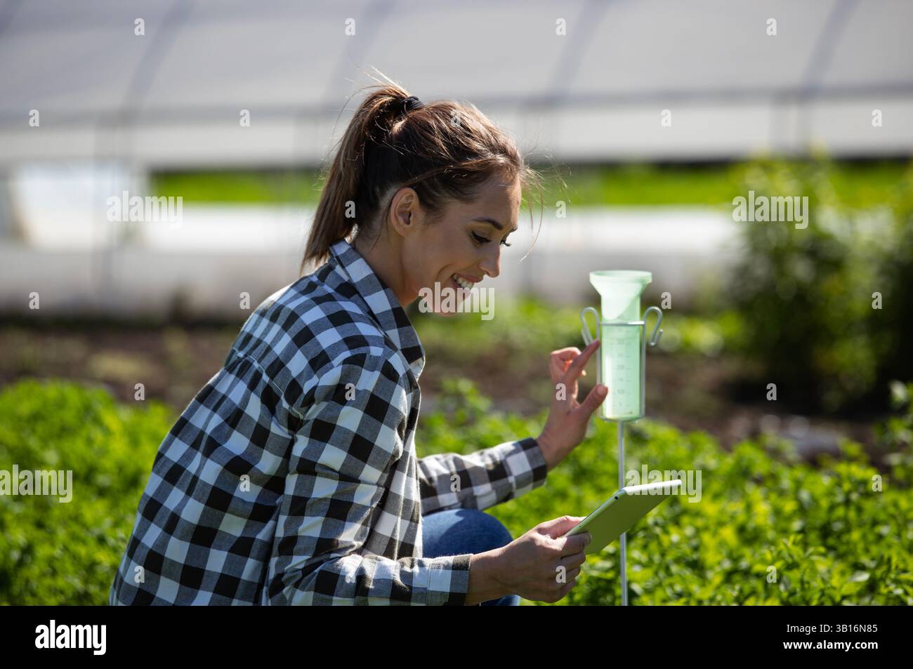 Donna che controlla i dati su un tablet mentre osserva il pluviometro e le piante in una lussureggiante serra. L'attività si svolge sotto un cielo limpido, mettendo in mostra le zone rurali Foto Stock