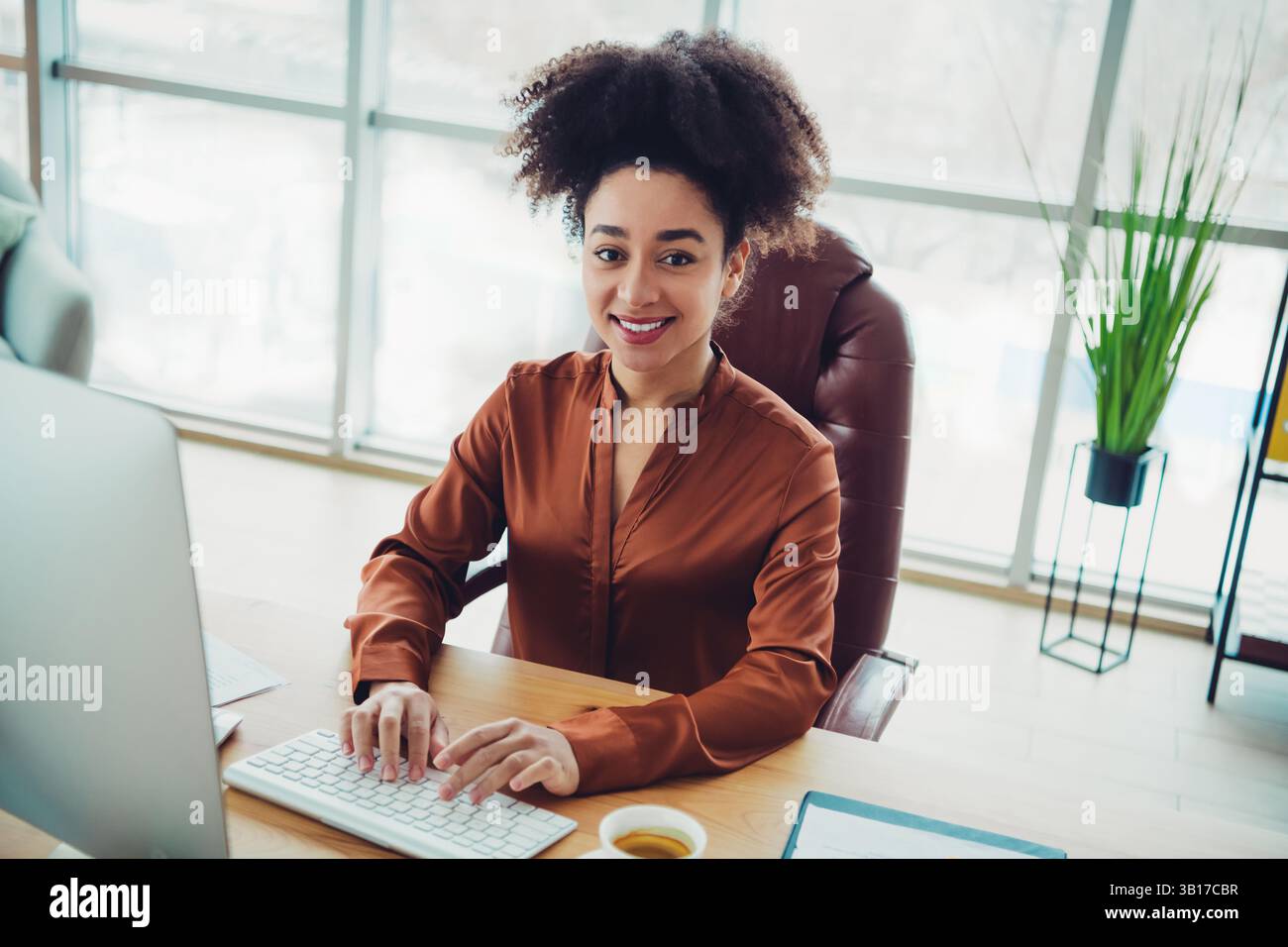 Giovane donna d'affari con capelli naturali alla sua scrivania in un ufficio moderno, sorridente e lavora al computer in piena luce naturale Foto Stock