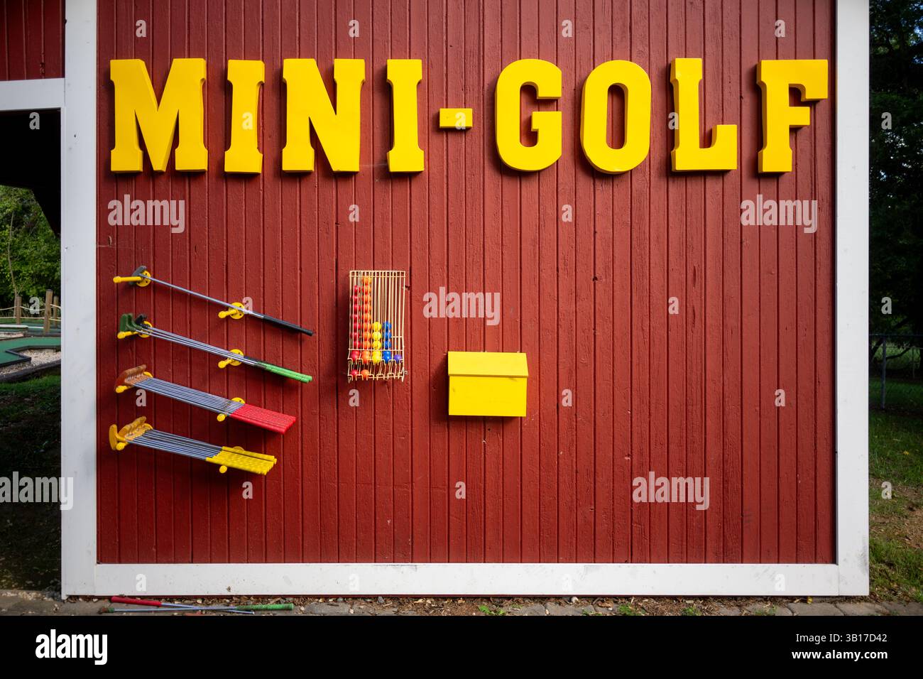 Insegna e attrezzatura da mini-golf Stati Uniti | STATI UNITI — Un muro di fienile rosso consumato porta un grande cartello giallo "Mini-Golf" sopra un espositore self-service di putter e un cestino di filo metallico con palline colorate in un campo da mini-golf lungo la strada. Le stazioni di attrezzatura self-service, comuni nei campi gestiti da famiglie dalla metà del ventesimo secolo, consentono ai giocatori di raccogliere bastoni e palline prima di una partita senza un bancone con personale. Foto Stock