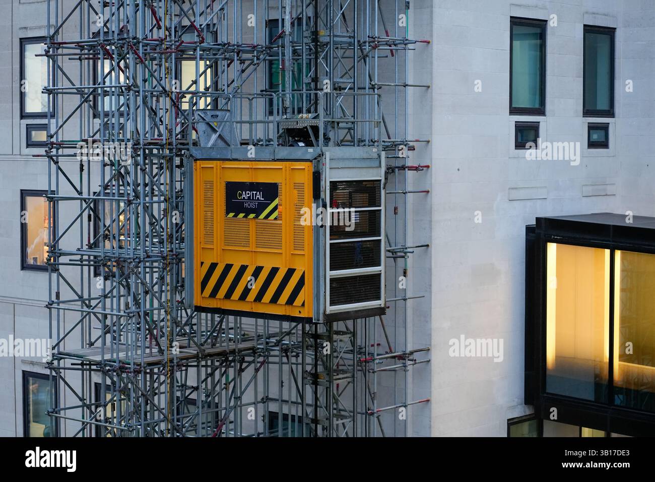 Un ascensore giallo per la costruzione del Capital Hoist è appeso su un'impalcatura contro un edificio moderno in un ambiente urbano. Foto Stock