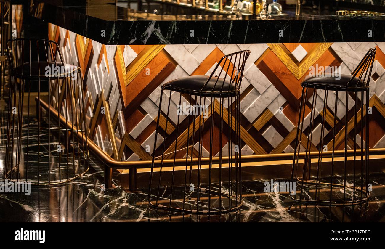 Elegante bancone bar con pannelli geometrici in legno e marmo, illuminato da una calda illuminazione, caratterizzato da moderni sgabelli in filo metallico e pavimento in marmo scuro. Foto Stock
