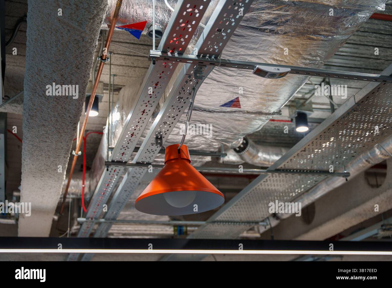 Una vivace lampada a sospensione industriale arancione è sospesa tra tubi esposti, condotti e passacavi nel soffitto di un edificio moderno. Foto Stock