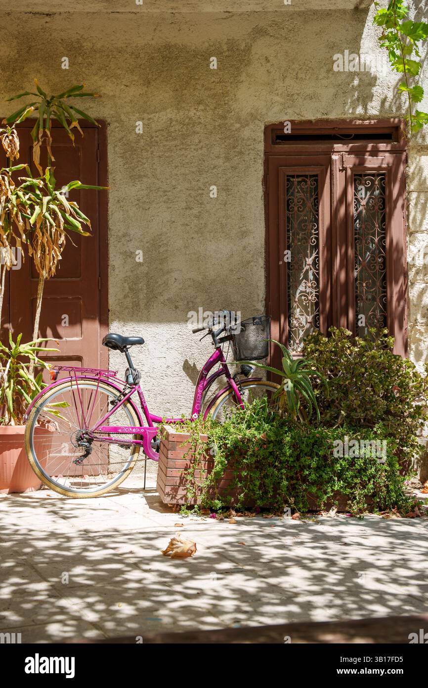 Una bicicletta viola si appoggia contro un muro illuminato dal sole all'esterno di una casa tradizionale con porte ornate e piante in vaso in una città di Creta, in Grecia. Foto Stock