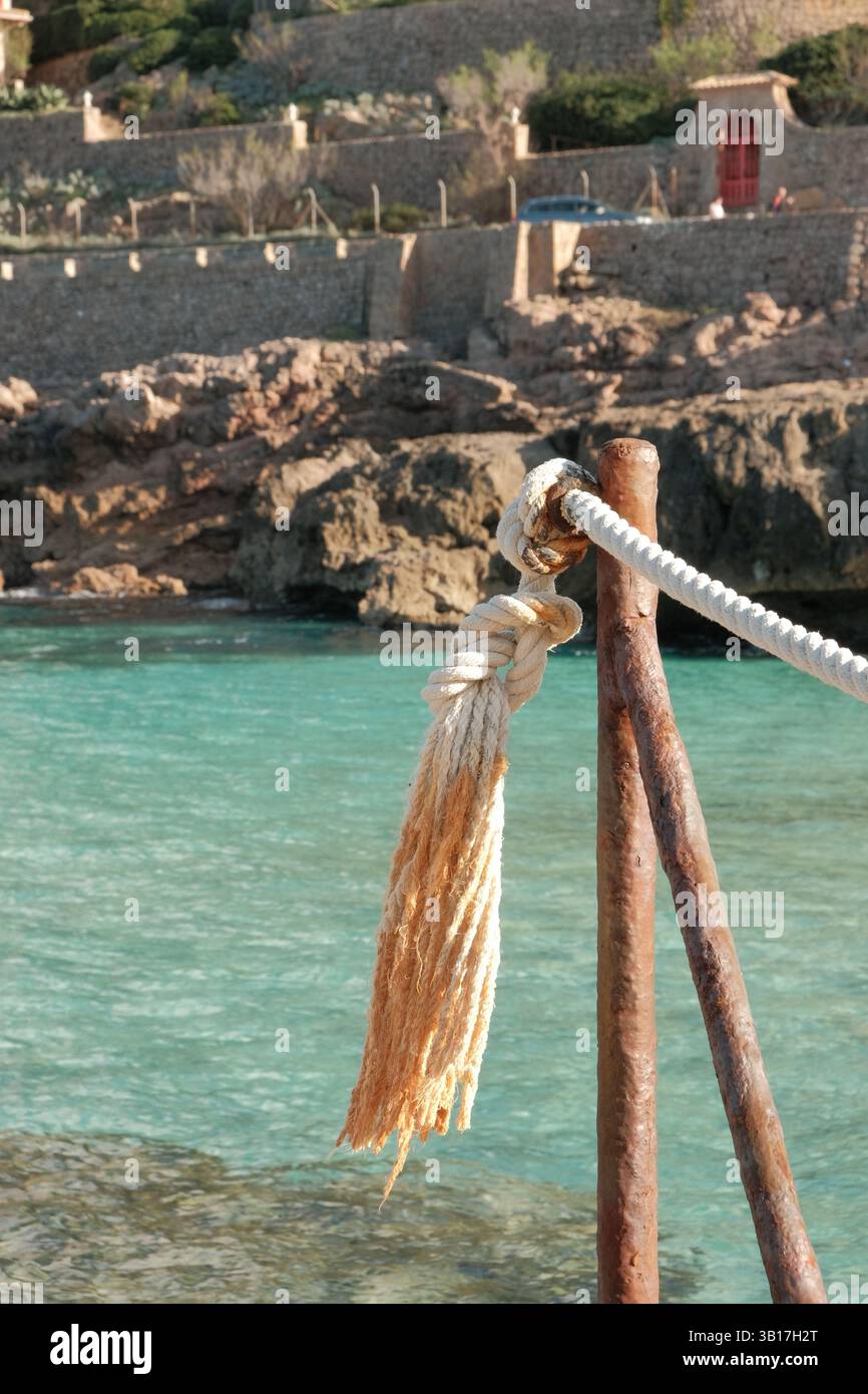 Ormeggio tradizionale di barche mediterranee con corda nautica e palo in legno indossato contro le acque turchesi e la costa rocciosa. Foto Stock
