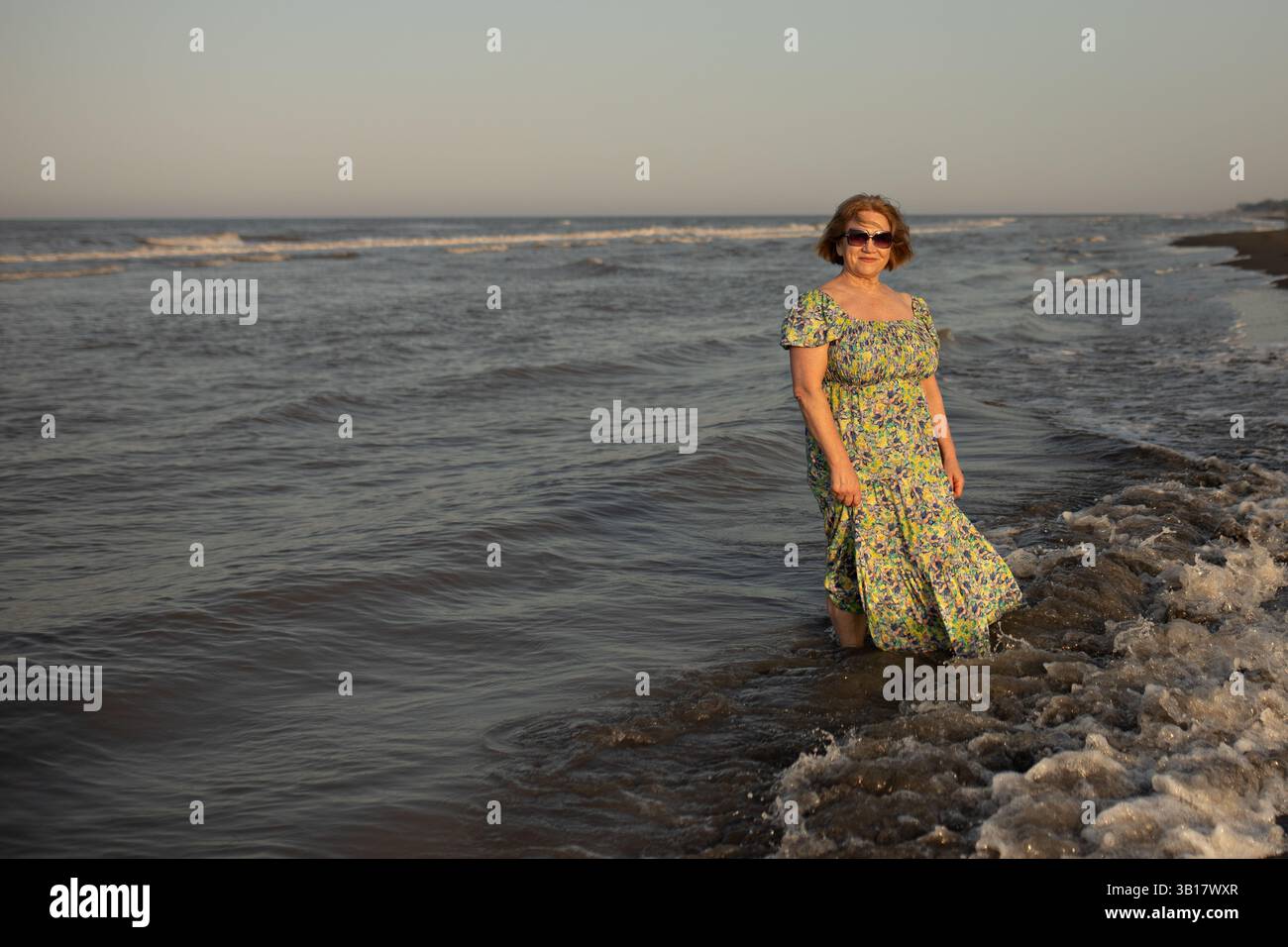Donna anziana in piedi al mare la sera. Indossa un maxi vestito floreale. L'atmosfera generale dell'immagine è tranquilla e serena, Foto Stock