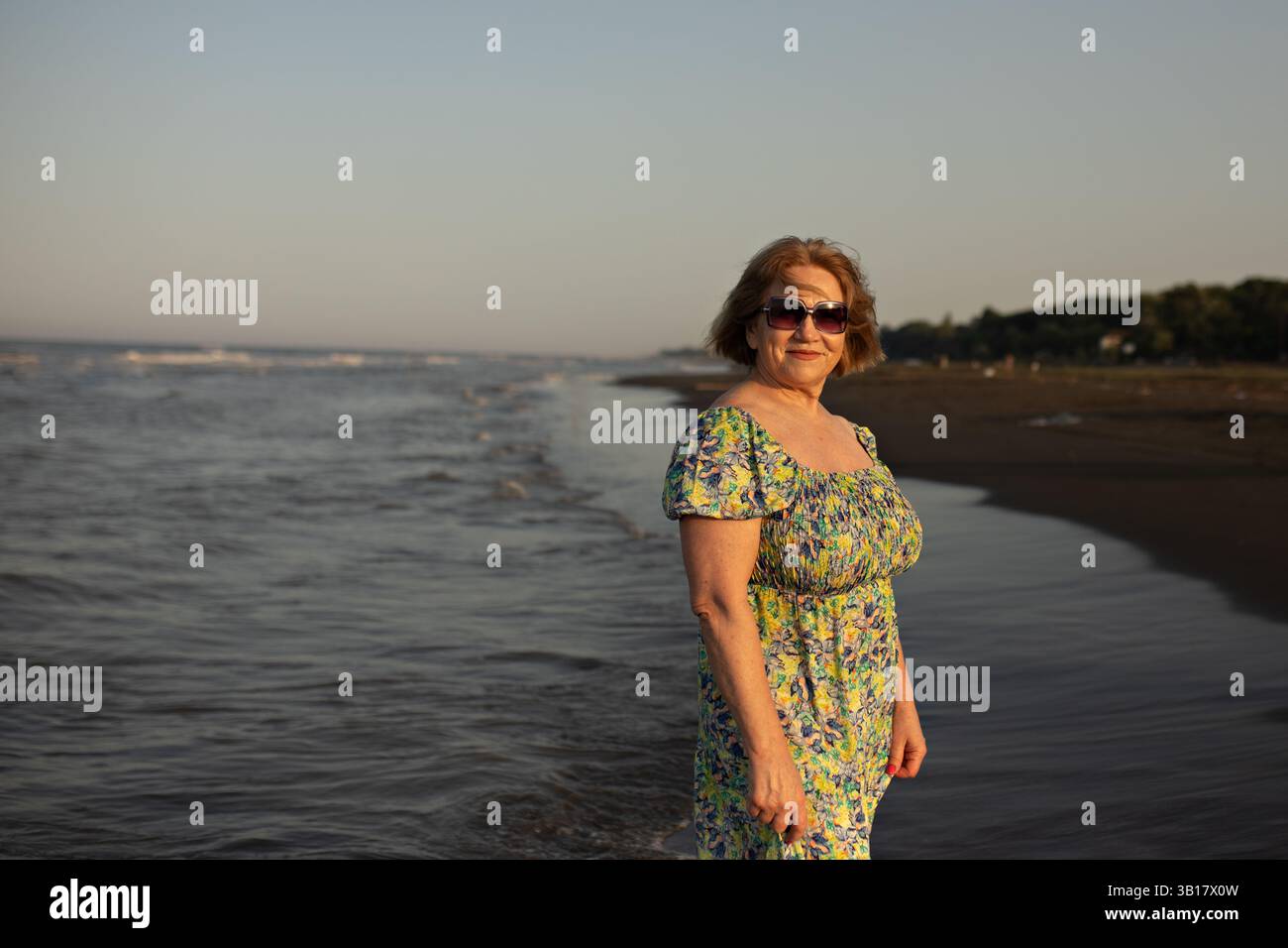 donna esterna in piedi al mare. Indossa un maxi abito floreale. La donna ha un'espressione calma e contenta Foto Stock