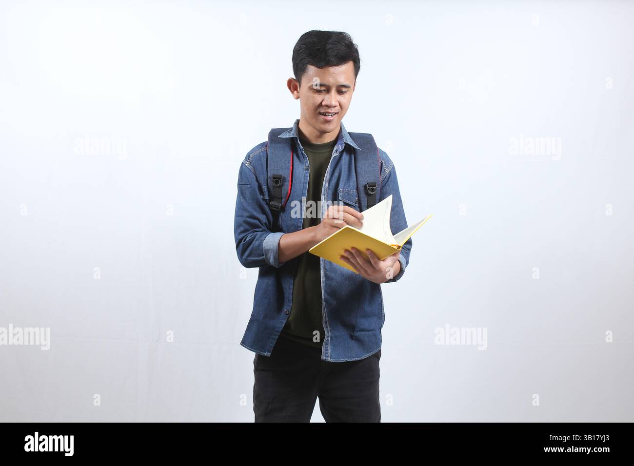 Uomo asiatico che indossa una camicia blu scuro in denim che porta zaino e libro aperto che studia per la preparazione all'esame, isolato su sfondo bianco Foto Stock