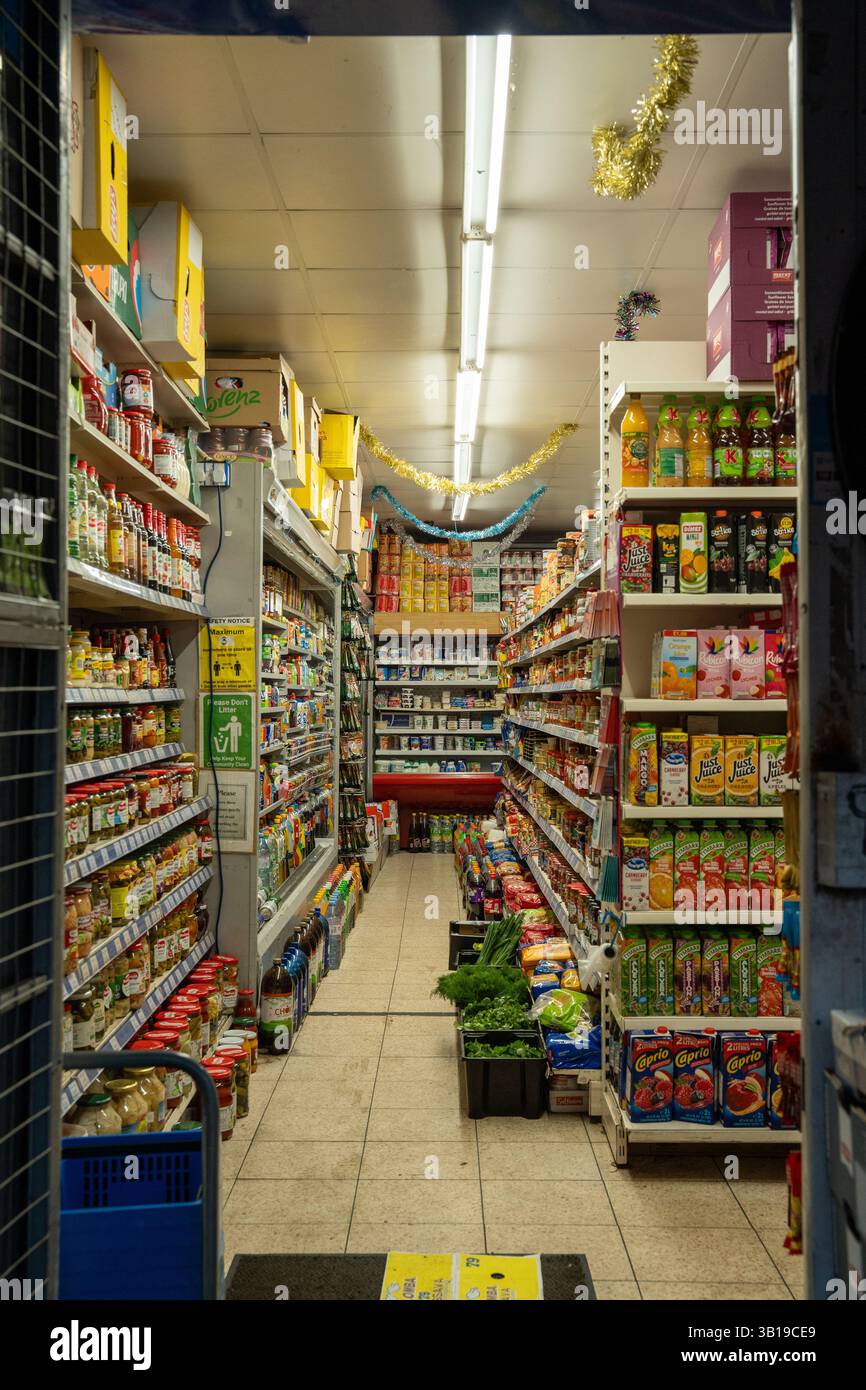 Corridoio stretto di un piccolo negozio di alimentari rifornito di vari prodotti alimentari, illuminato da luci fluorescenti, decorazioni festose appese al ceil Foto Stock
