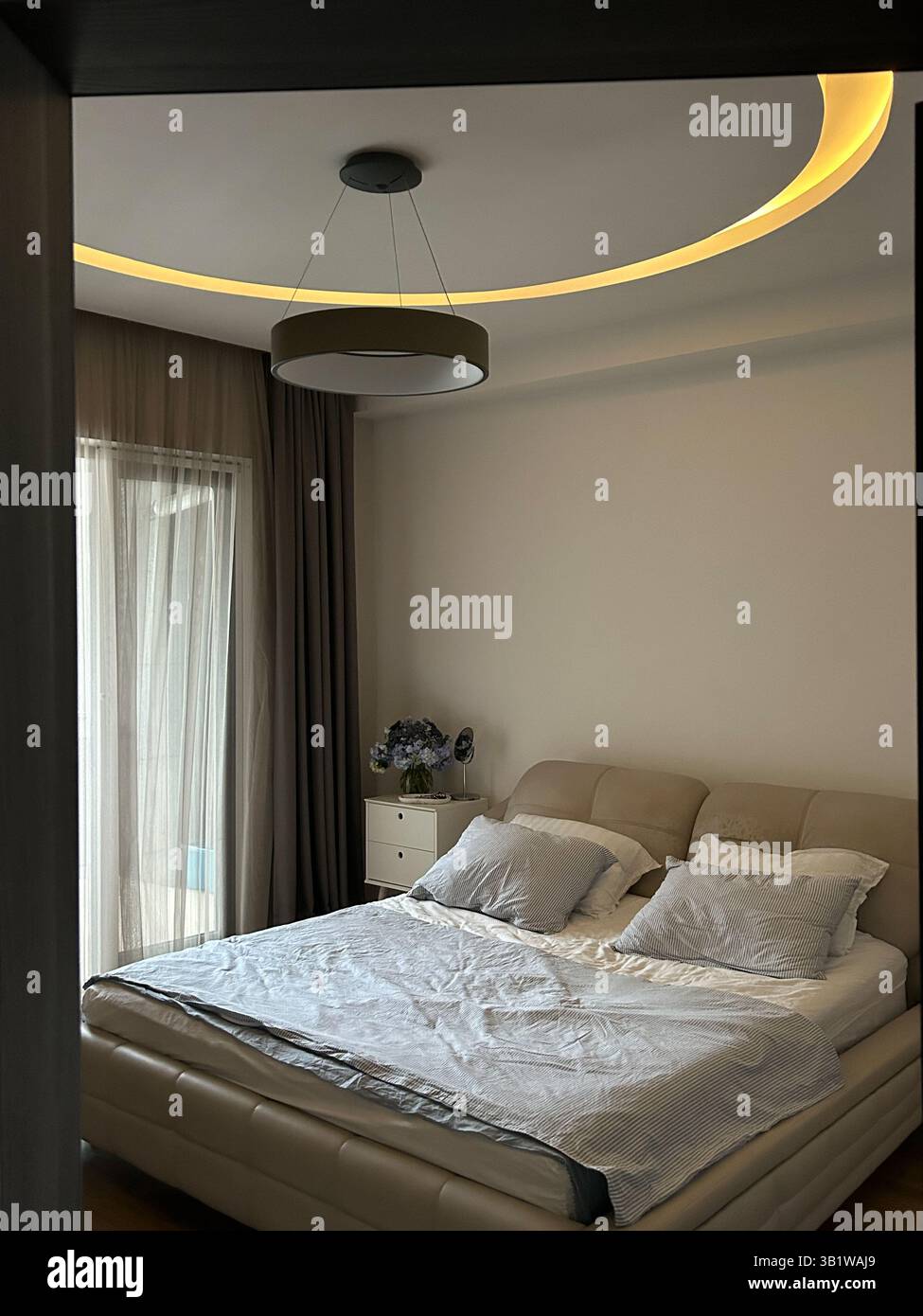 La foto mostra una camera da letto moderna con un letto matrimoniale imbottito beige, biancheria da letto leggera e numerosi cuscini. Una plafoniera rotonda con un LED circolare A. Foto Stock