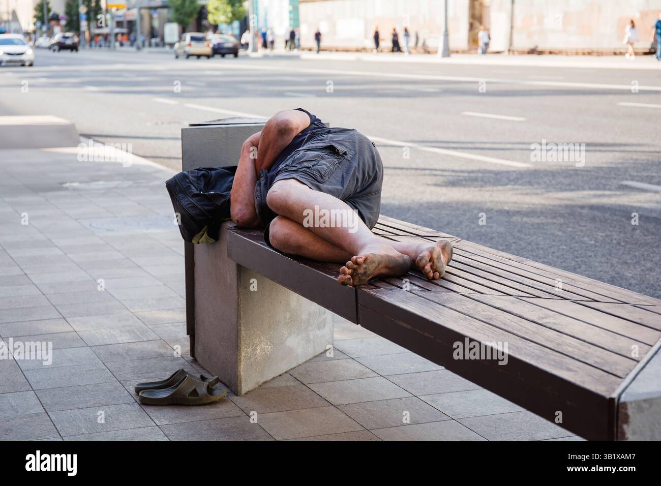 Un senzatetto irriconoscibile a piedi nudi dorme su una panchina di una strada cittadina, senza tetto, problemi sociali Foto Stock