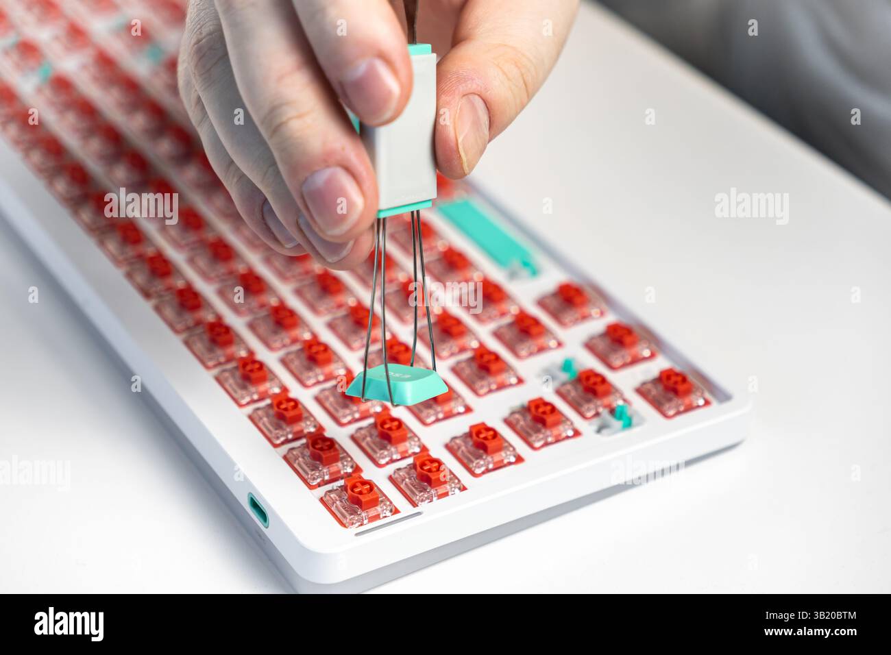 Primo piano di una mano che tiene in mano un estrattore per tappi a chiave per rimuovere un tasto color verde acqua da una tastiera meccanica con interruttori rossi, evidenziando una personalizzazione fai da te pr Foto Stock