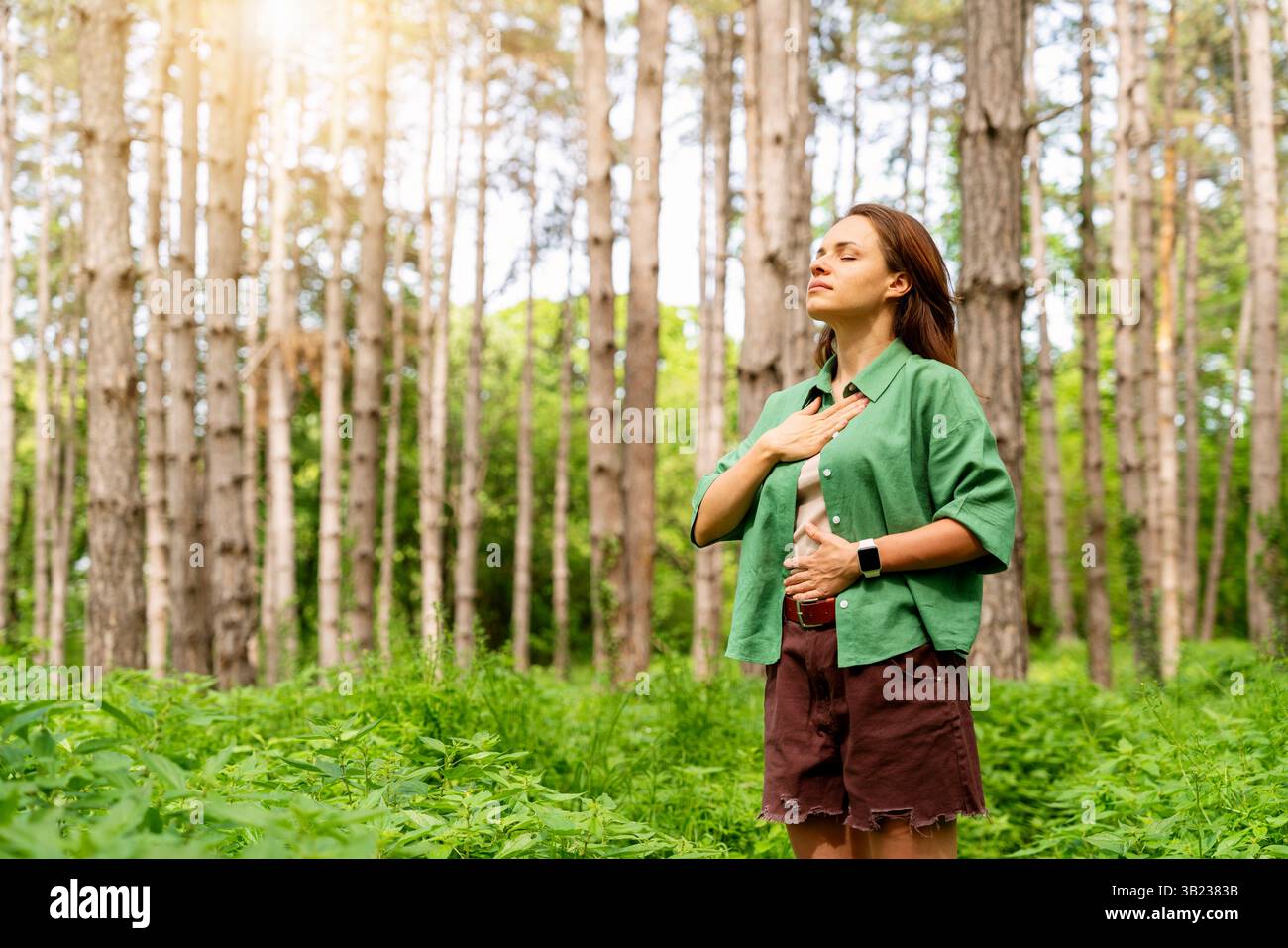 Giovane donna adulta in camicia verde che pratica esercizio respiratorio nella foresta. Foto Stock