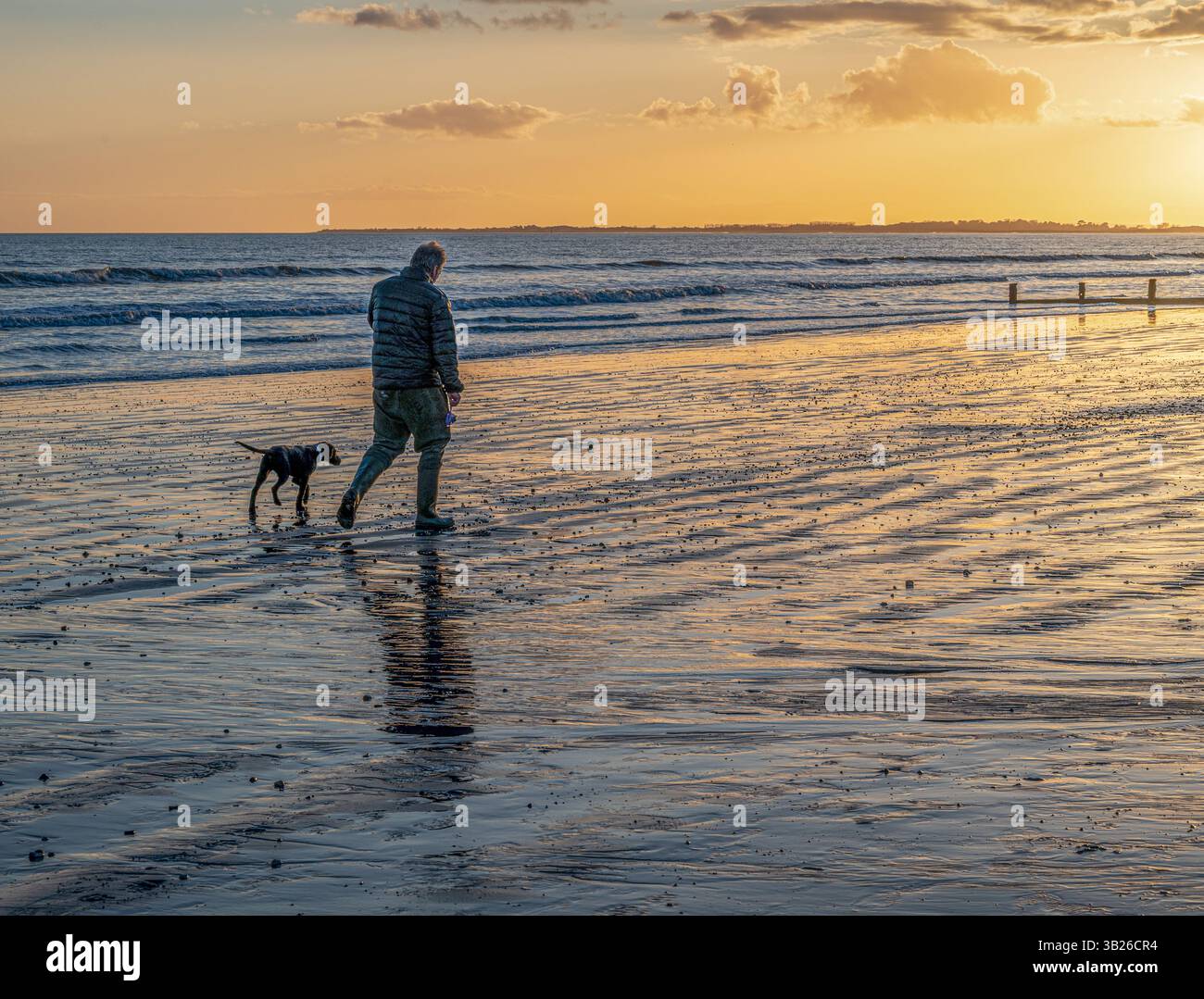 Un uomo solitario che cammina il suo cane lungo una spiaggia con la bassa marea e il tramonto. Foto Stock
