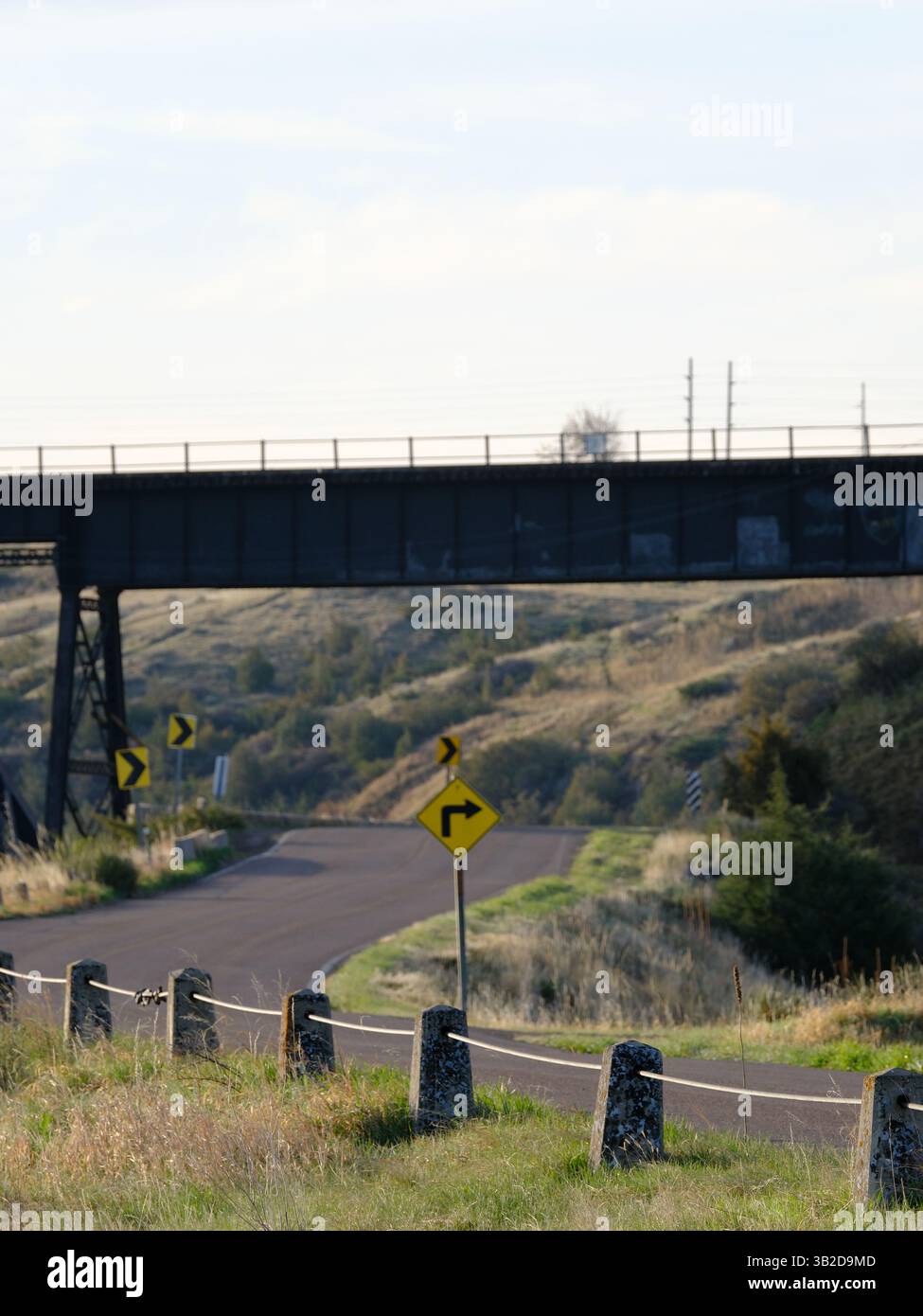 Strada curva sotto il ponte ferroviario - Una tranquilla strada rurale si snoda sotto un ponte ferroviario in acciaio, contrassegnato da indicazioni gialle Foto Stock