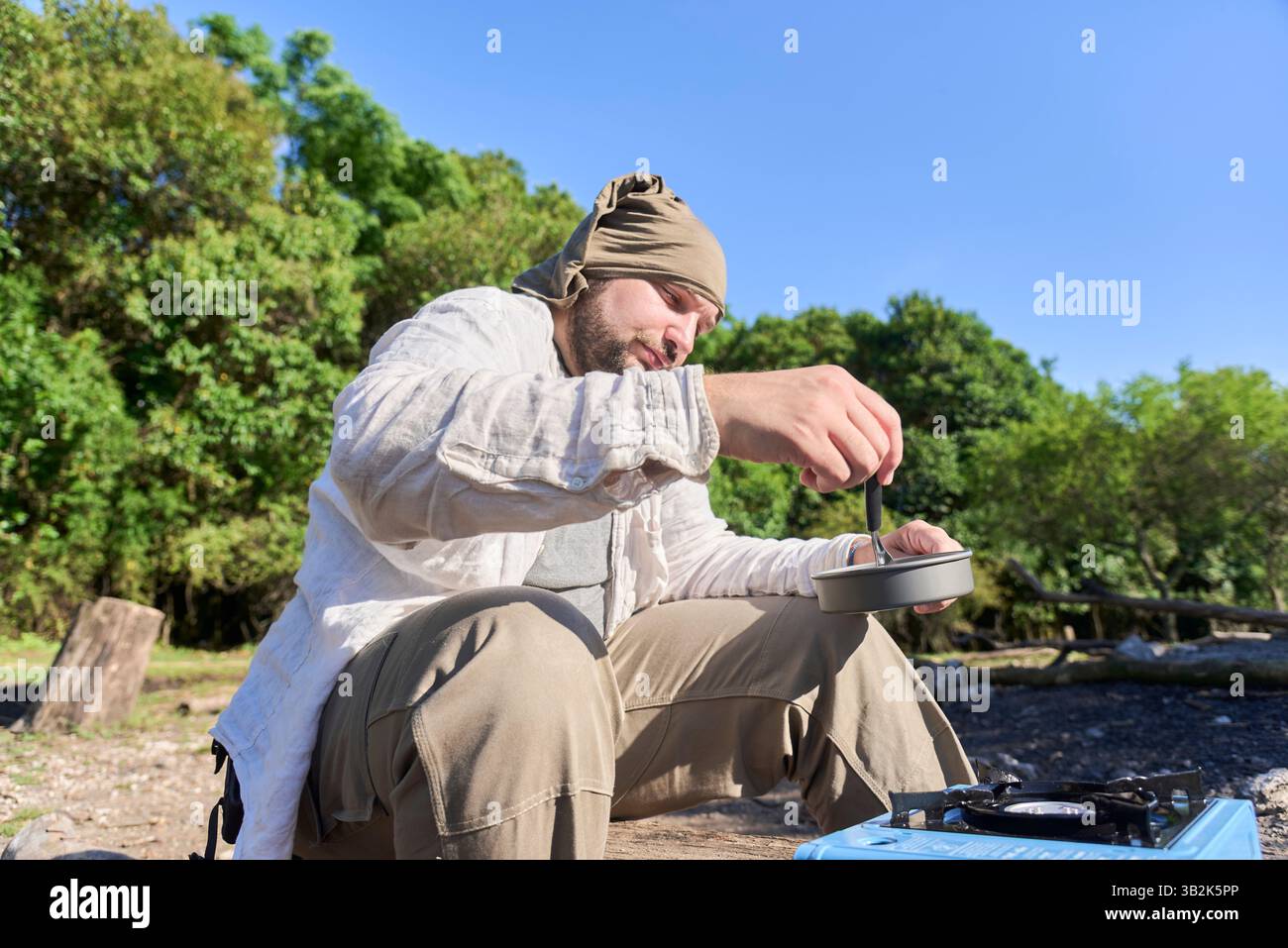 Viaggiatore ispanico che cucina all'aperto su una stufa portatile azzurra mentre campeggia in un campeggio naturale una mattina di sole. Foto Stock