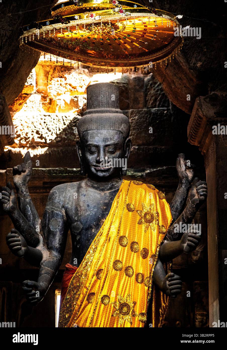 Una maestosa statua di una divinità adornata con una veste dorata e circondata da intricate incisioni in pietra. La figura ha più braccia e un sereno espresso Foto Stock