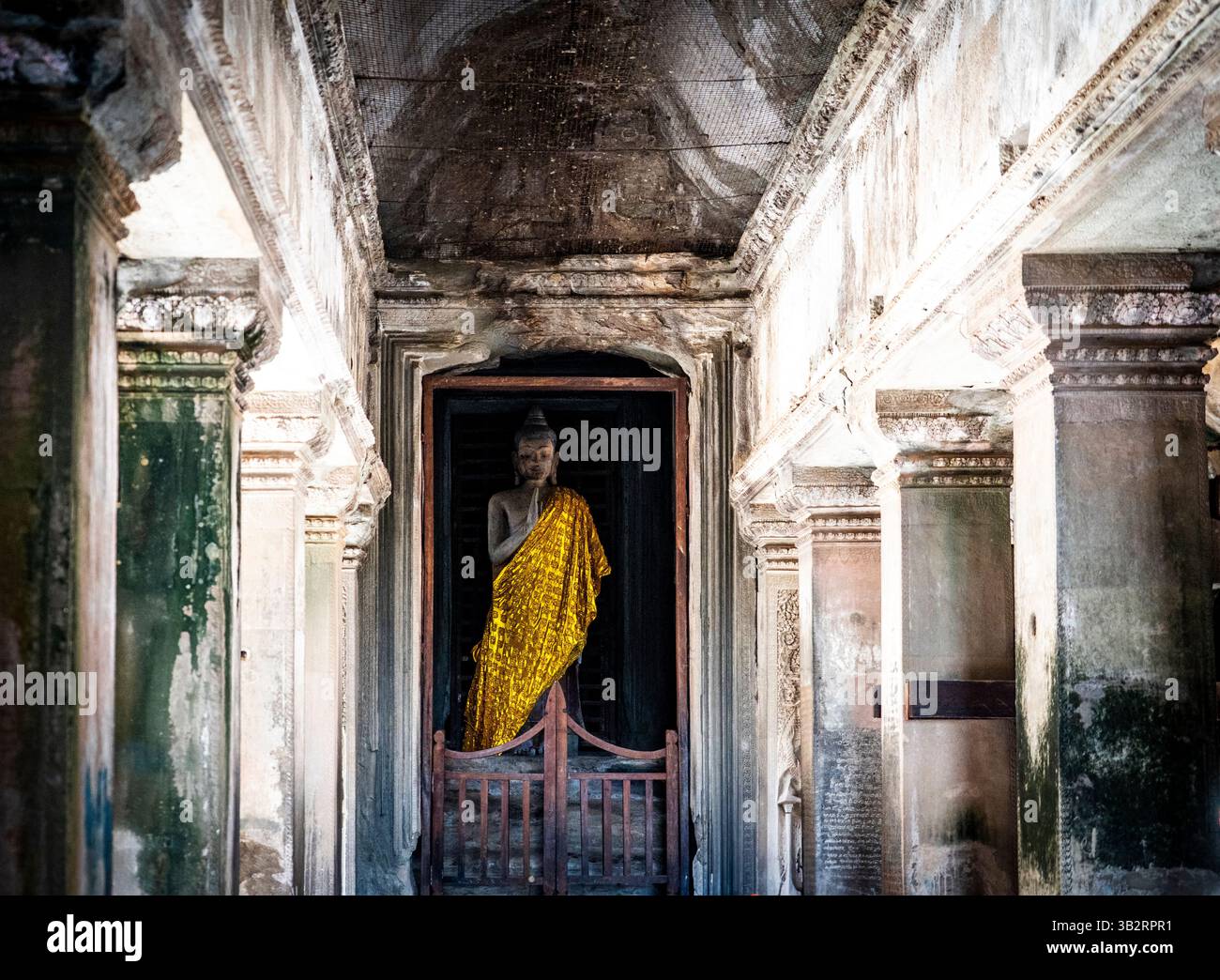 Una tranquilla statua di Buddha drappeggiata in una veste dorata sorge in un corridoio poco illuminato di un antico tempio, circondato da colonne di pietra e intri intemprati Foto Stock