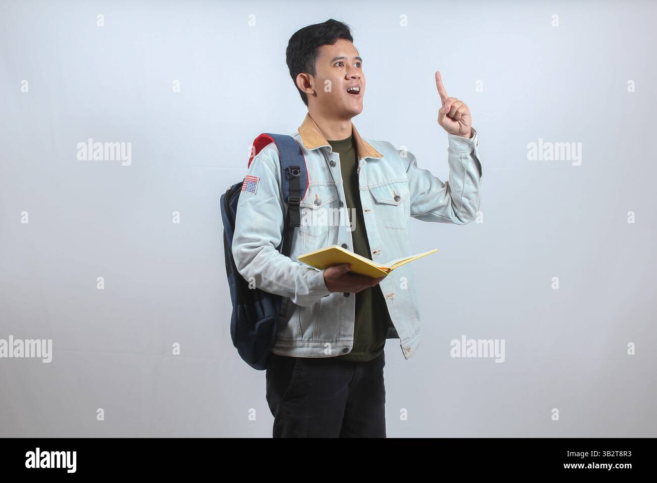 Uomo asiatico che indossa una giacca di denim che trasporta libri e borsa mostrando un dito su di un'idea brillante isolata su sfondo bianco, concetto educativo Foto Stock
