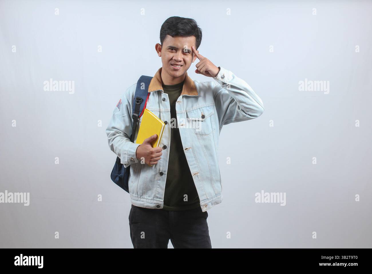 Uomo asiatico che indossa una giacca di denim con borsa e un libro che dà il segno di pensare seriamente puntando il dito verso la testa segnalando di pensare duro Foto Stock