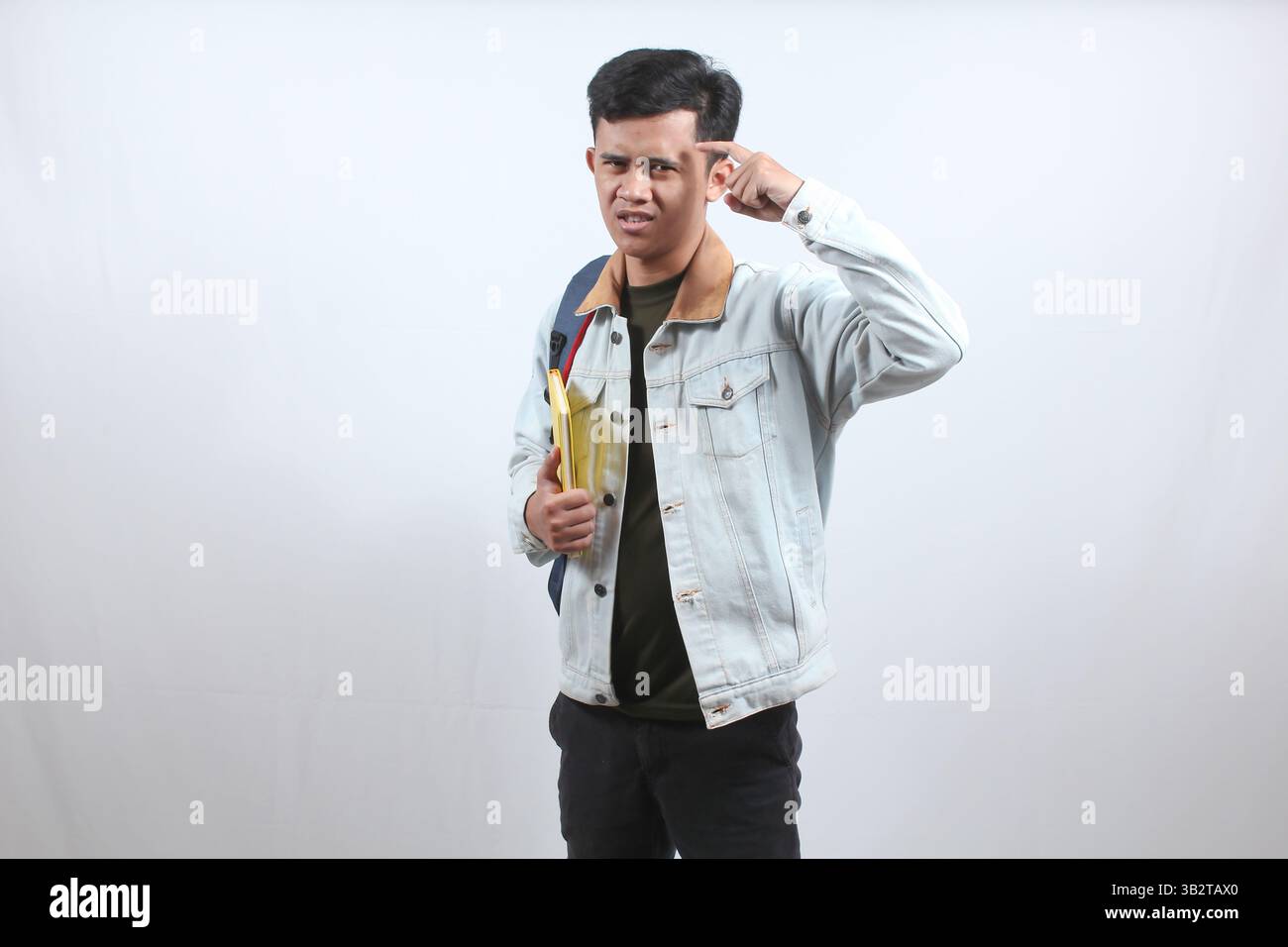 Uomo asiatico che indossa una giacca di denim con borsa e un libro che dà il segno di pensare seriamente puntando il dito verso la testa segnalando di pensare duro Foto Stock