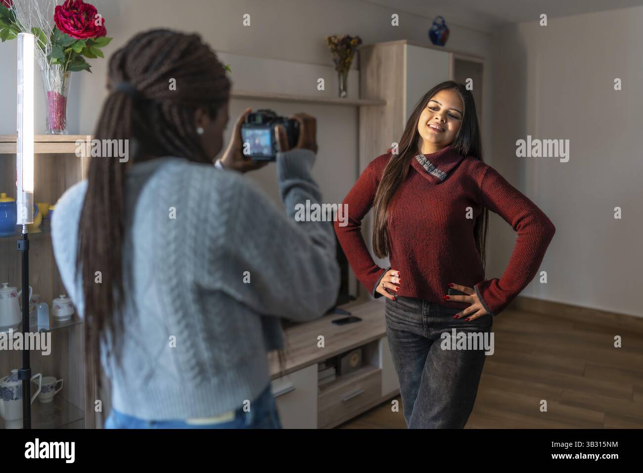 Giovane fotografo che cattura i momenti eleganti di una modella in posa a casa, utilizzando una luce LED brillante per un tocco creativo Foto Stock