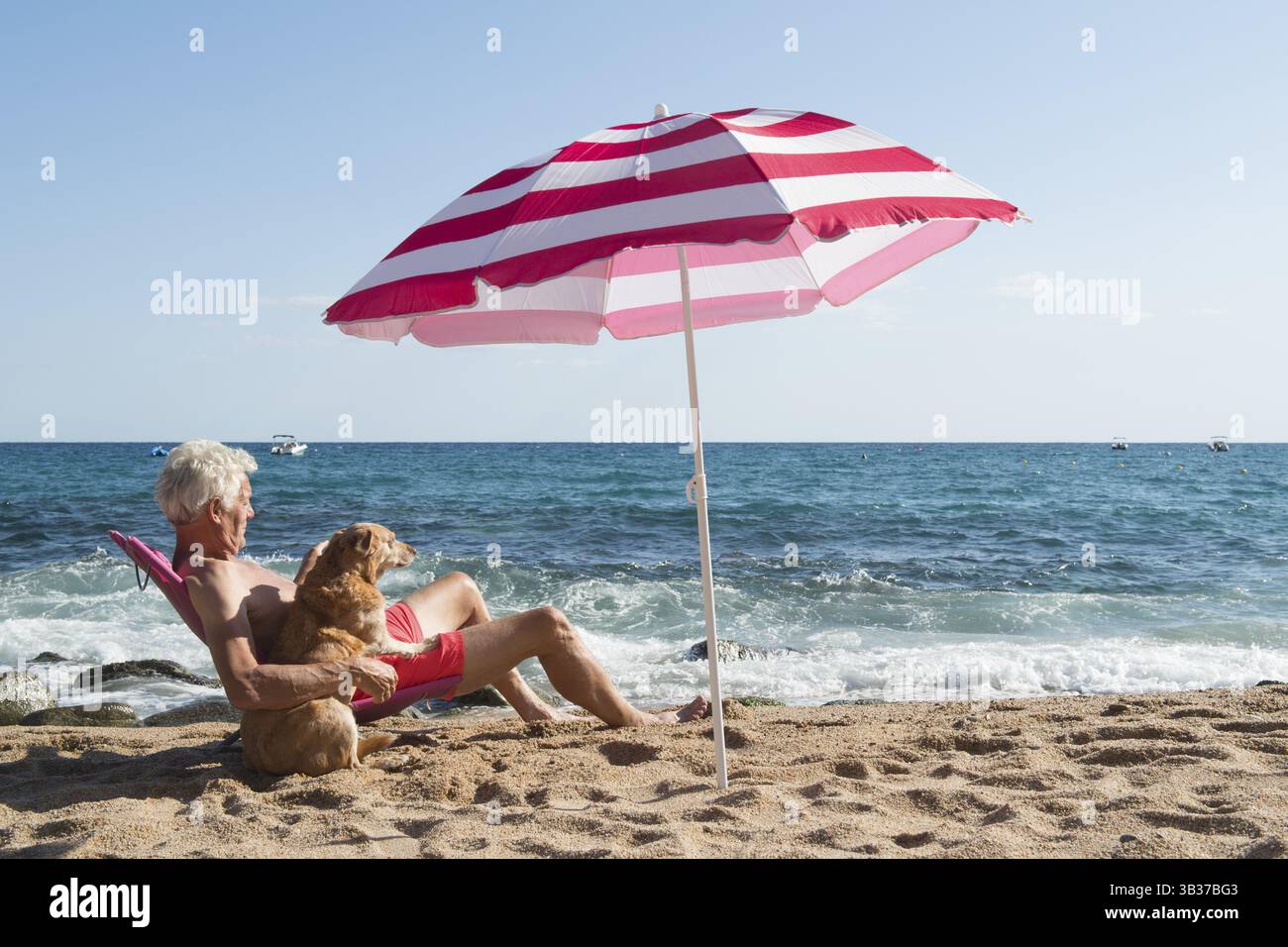 L'anziano prende il sole con il suo cane in spiaggia sotto l'ombrellone con onde, mare e sabbia Foto Stock