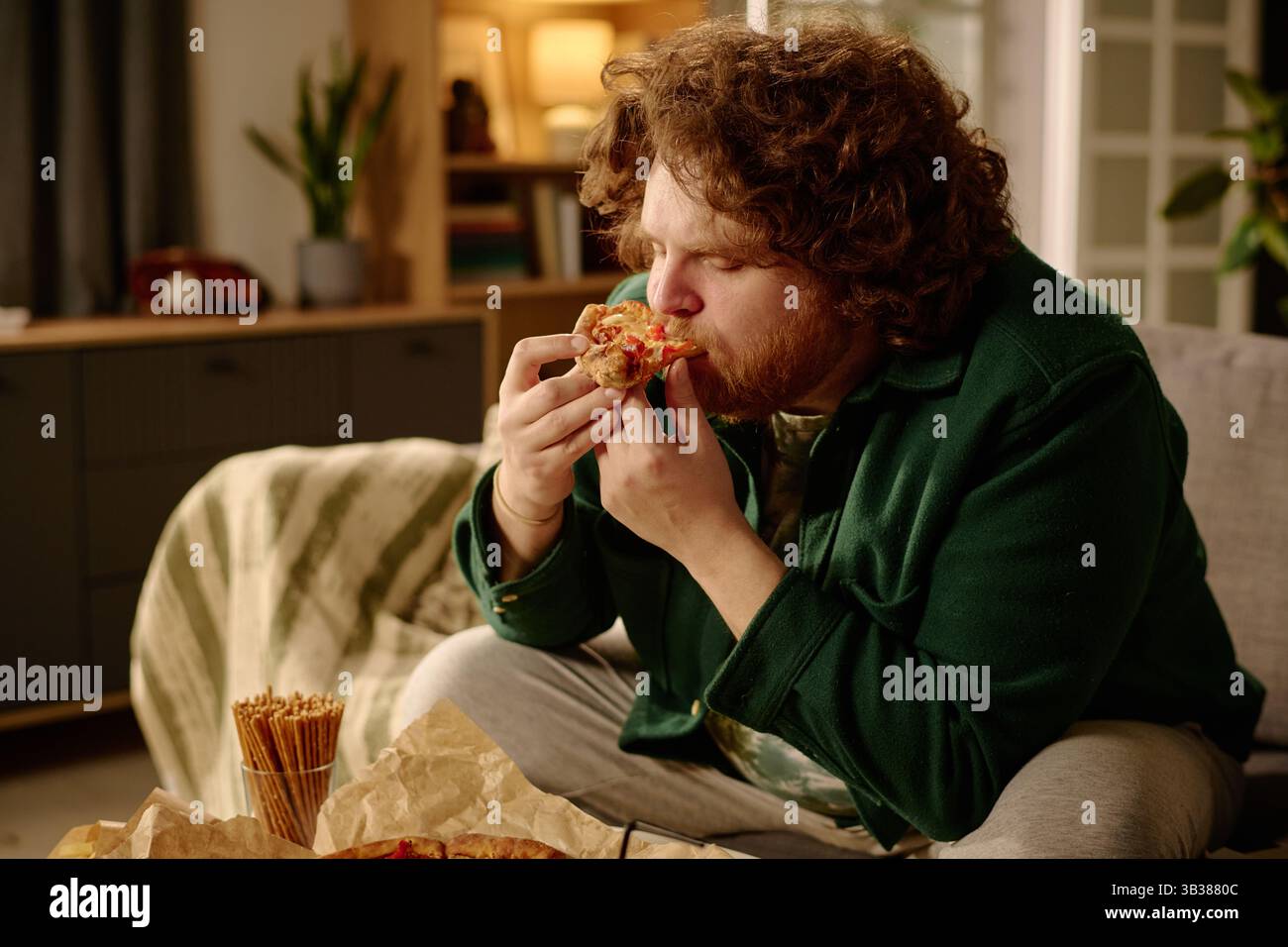 L'uomo barbuto seduto su un divano sta gustando una fetta di pizza in un accogliente salotto con una luce calda. Lo sfondo include un divano, elementi decorativi e una libreria parzialmente visibile Foto Stock