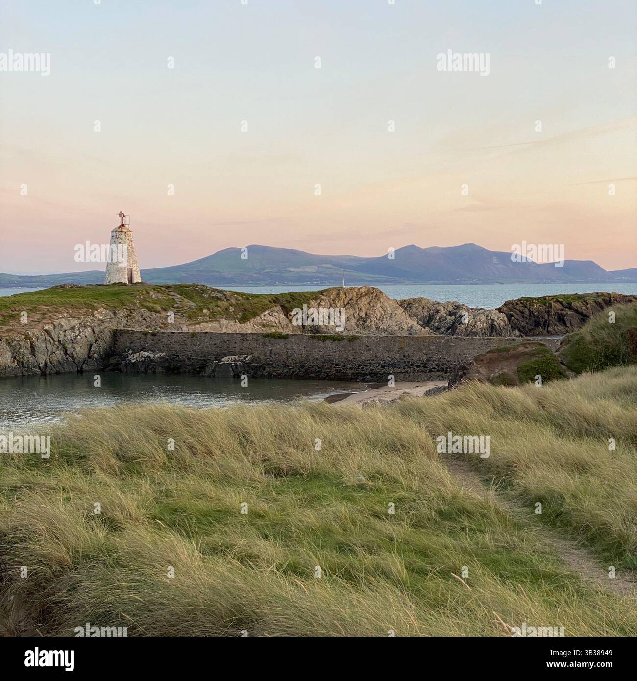 Una splendida foto di un faro e di una baia a Ynys Llandwyn al tramonto, con il cielo rosa e le viste mozzafiato di Snowdonia sullo sfondo - Immagine stock catturata con smartphone