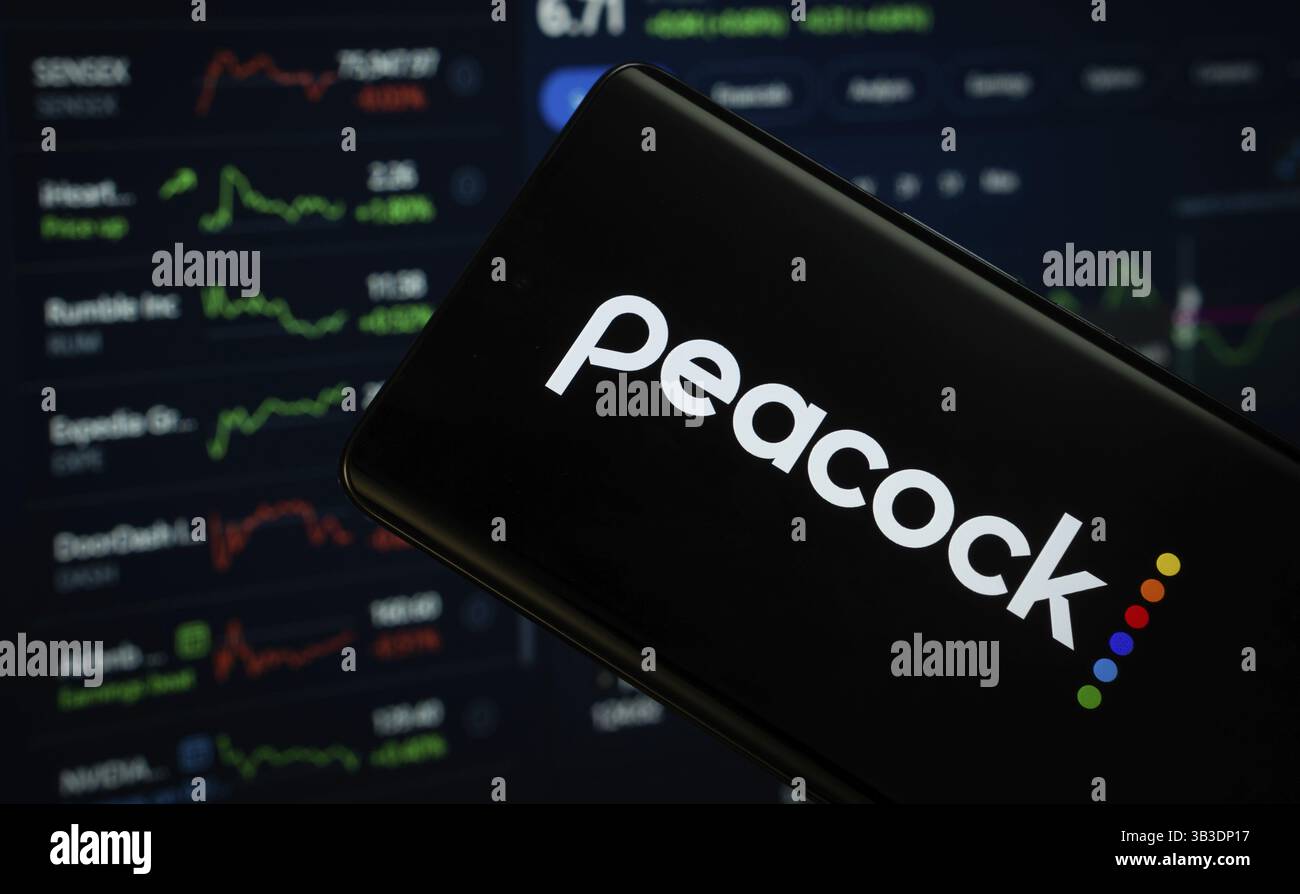 Dhaka, Bangladesh - 17 marzo 2025: Il logo peacock viene visualizzato sullo schermo di uno smartphone, con una pagina Web del grafico delle scorte visibile sullo schermo di un computer nel bac Foto Stock