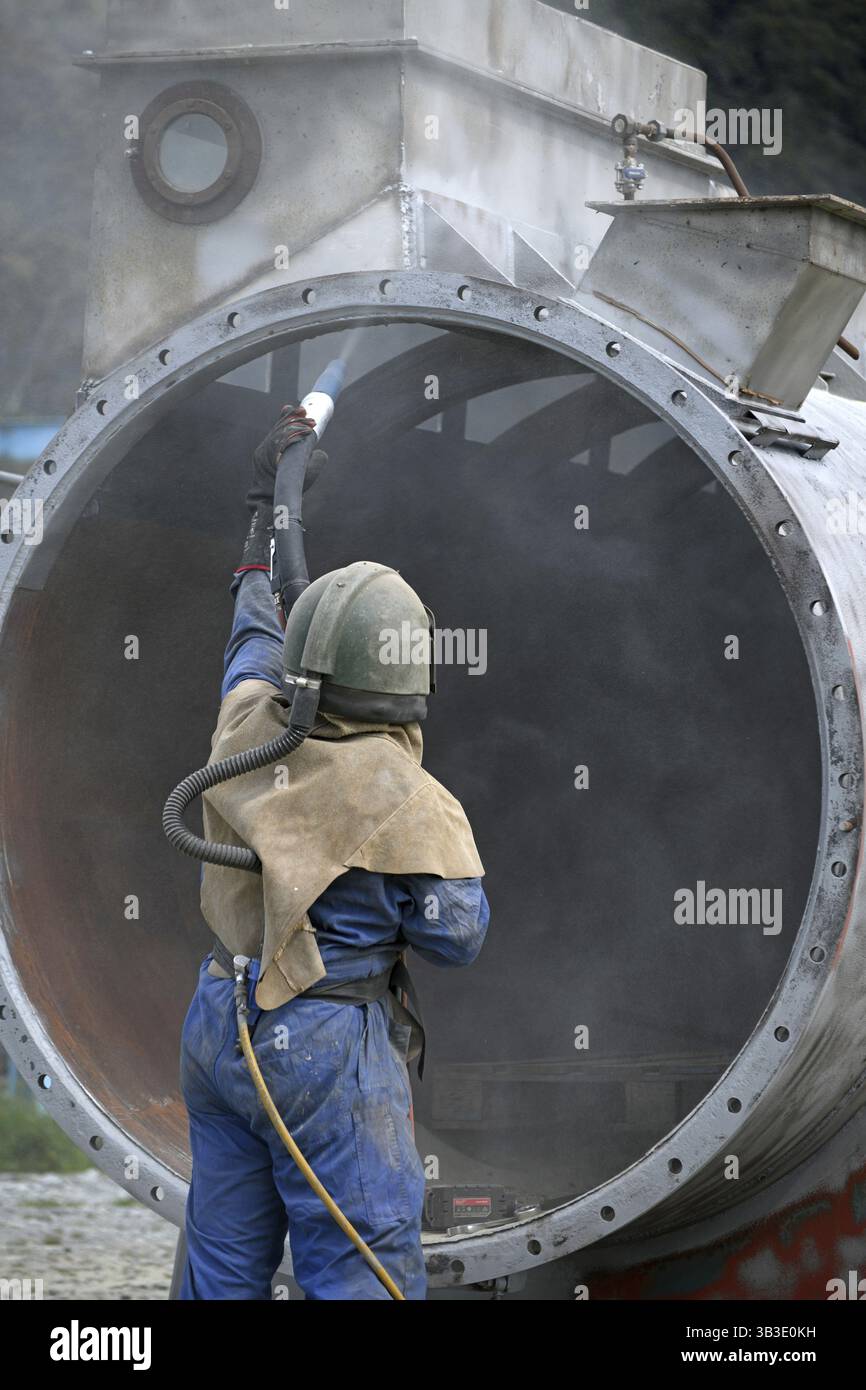 Un uomo che indossa l'attrezzatura di sicurezza completa utilizza vetro macinato per sabbiare il rivestimento in acciaio di una macchina per l'essiccazione della carne per un impianto di rendering, West Coast, nuova Zelanda Foto Stock