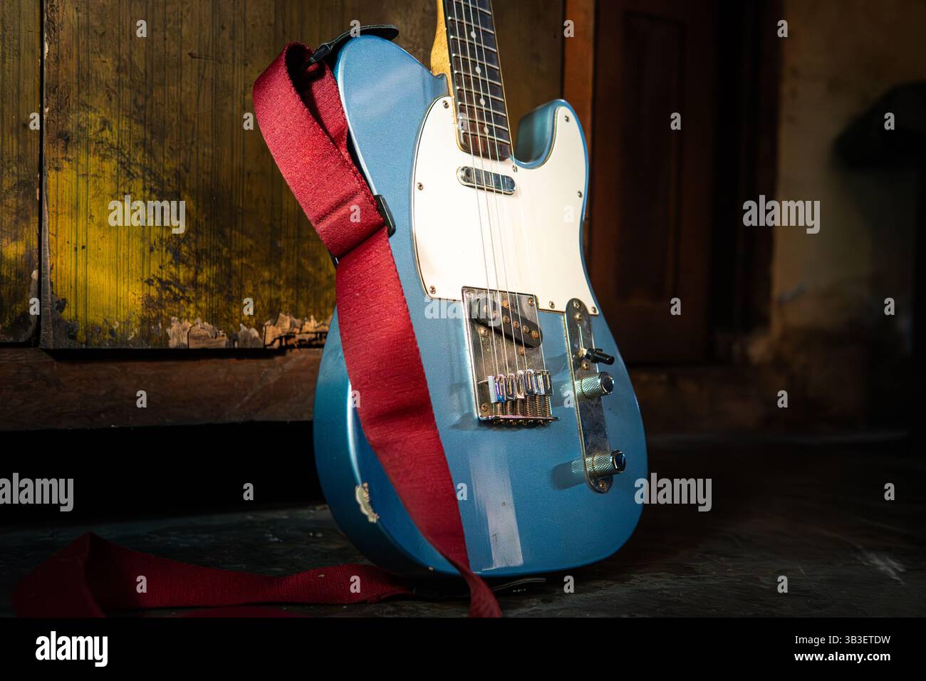 Chitarra elettrica blu con cinturino rosso appoggiato sul pavimento, inondata di luce naturale. Ideale per musica, stile di vita o progetti a tema vintage. Foto Stock