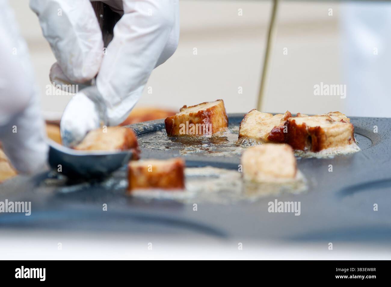 Frittura di salsicce a fette di olio su una piastra calda, maneggiata con un attrezzo metallico con guanti durante la preparazione professionale degli alimenti. Foto Stock