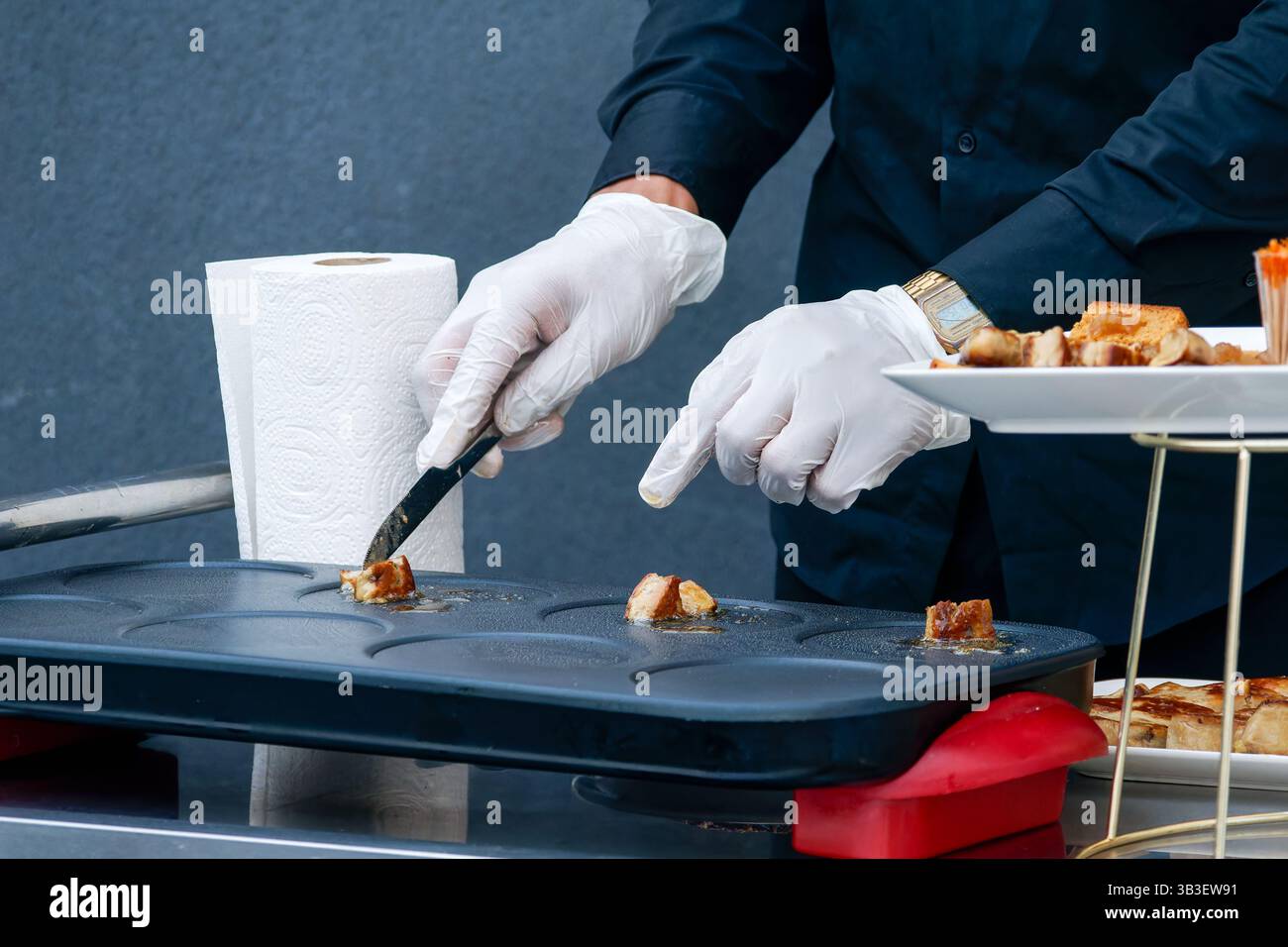Frittura di salsicce a fette di olio su una piastra calda, maneggiata con un attrezzo metallico con guanti durante la preparazione professionale degli alimenti. Foto Stock
