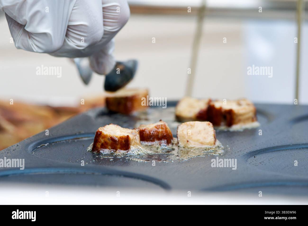 Frittura di salsicce a fette di olio su una piastra calda, maneggiata con un attrezzo metallico con guanti durante la preparazione professionale degli alimenti. Foto Stock