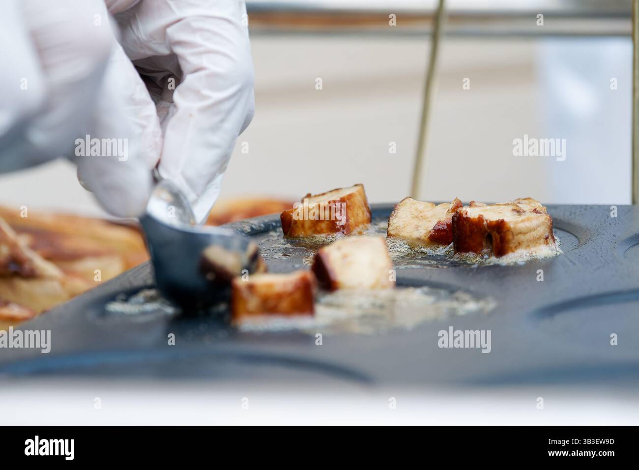 Frittura di salsicce a fette di olio su una piastra calda, maneggiata con un attrezzo metallico con guanti durante la preparazione professionale degli alimenti. Foto Stock