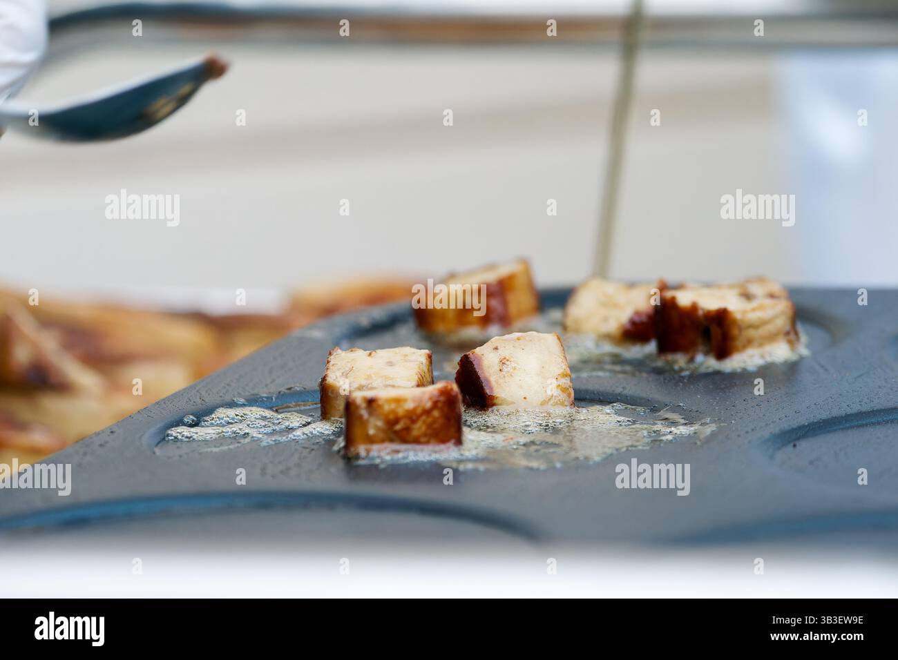 Frittura di salsicce a fette di olio su una piastra calda, maneggiata con un attrezzo metallico con guanti durante la preparazione professionale degli alimenti. Foto Stock